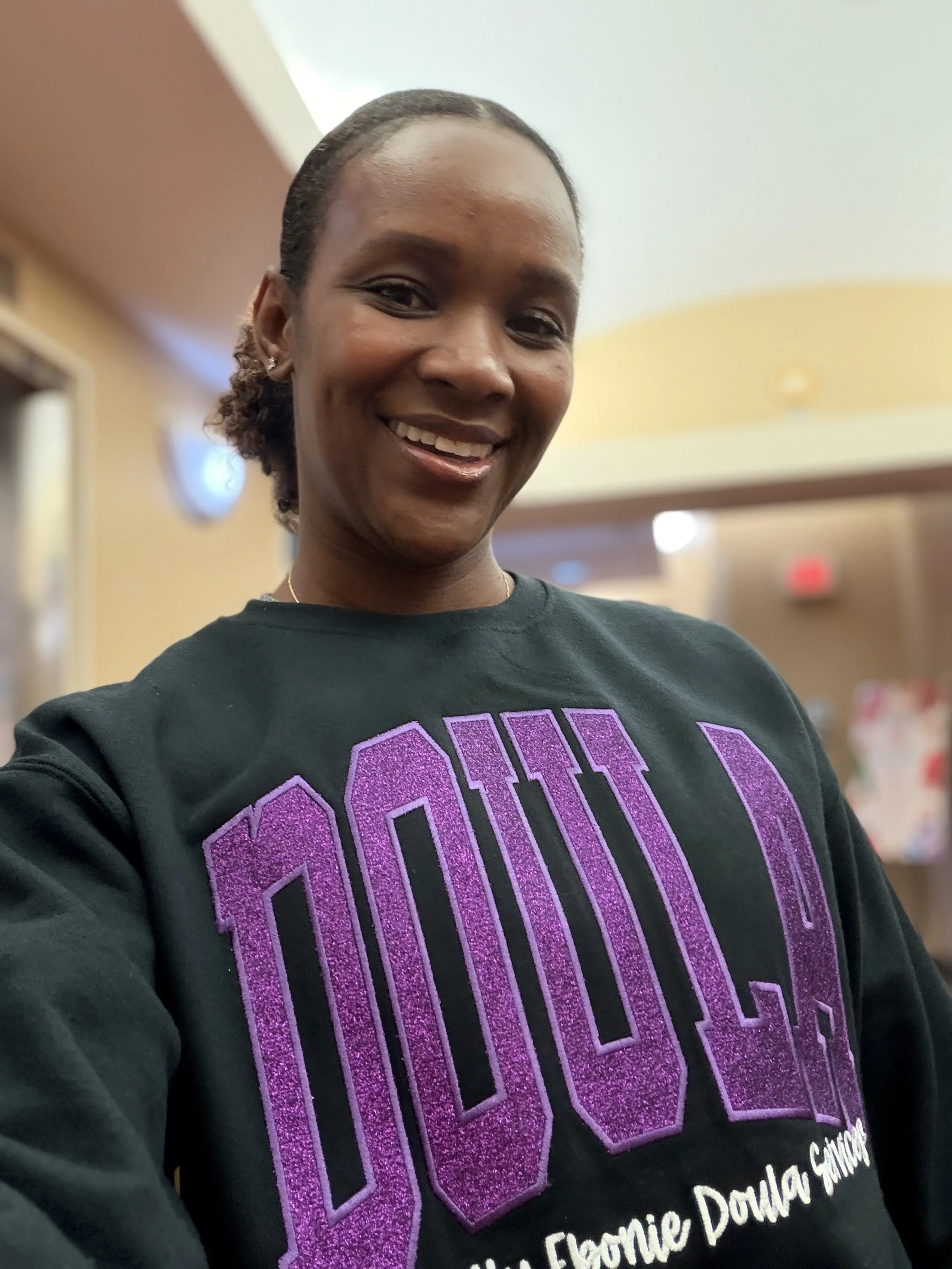 Close-up of a smiling woman with black hair, wearing earrings, and a black sweatshirt with purple glittery text that says 'OCA' and smaller white text underneath. The background appears to be an indoor setting with warm lighting.