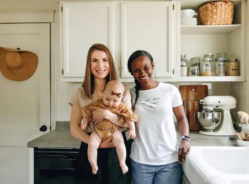 Two women and a baby in a kitchen. One woman is holding the baby, and both women are smiling. Ebonie Bailey on the right supporting one of her birth clients.