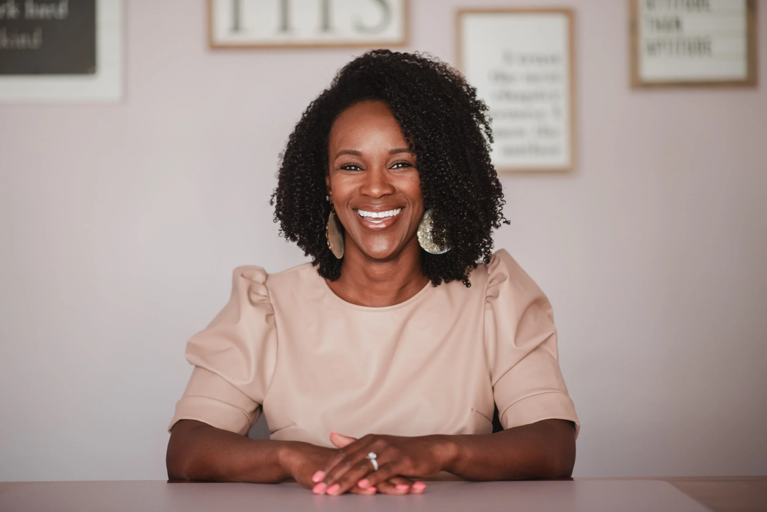 Smiling woman with curly hair wearing a beige top, sitting with hands clasped on a table, against a light-colored wall with framed posters.