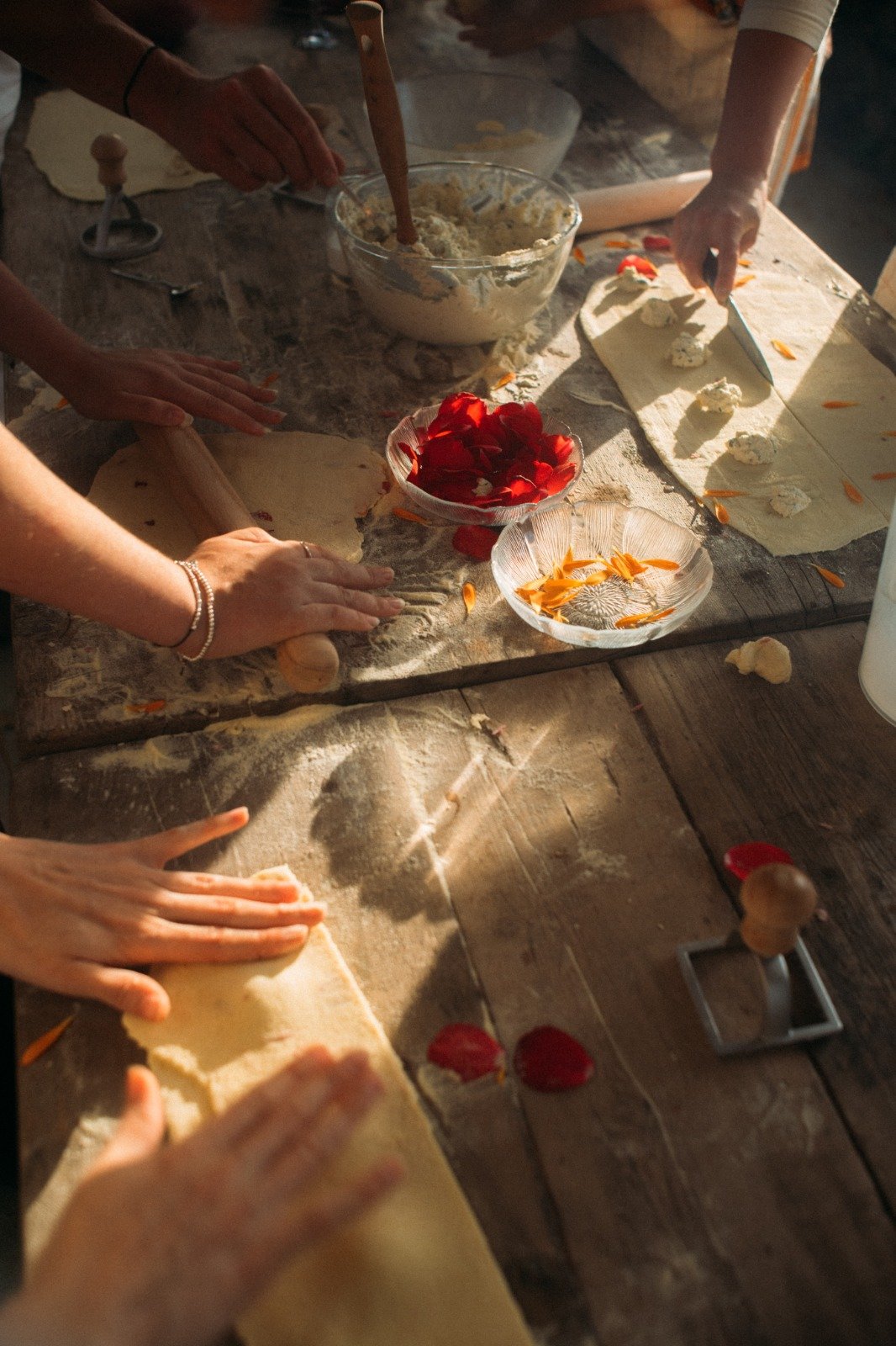 People making pasta on a rustic wooden table, using rolling pins to flatten dough. Bowls of red and orange flower petals are nearby. Ingredients for pasta filling are placed on dough sheets.