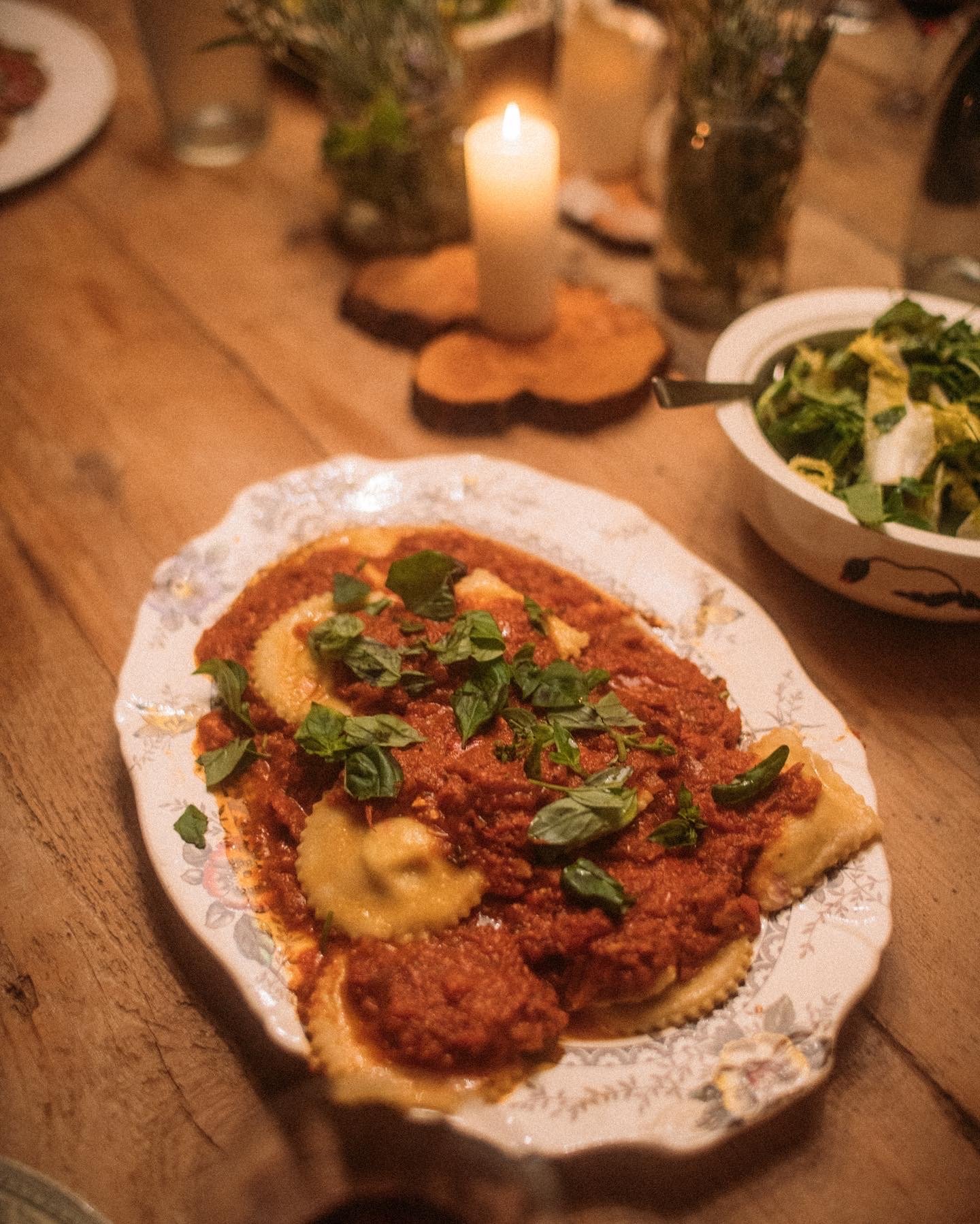 A plate of ravioli topped with tomato sauce and basil on a wooden table, with a candle and a bowl of salad in the background.