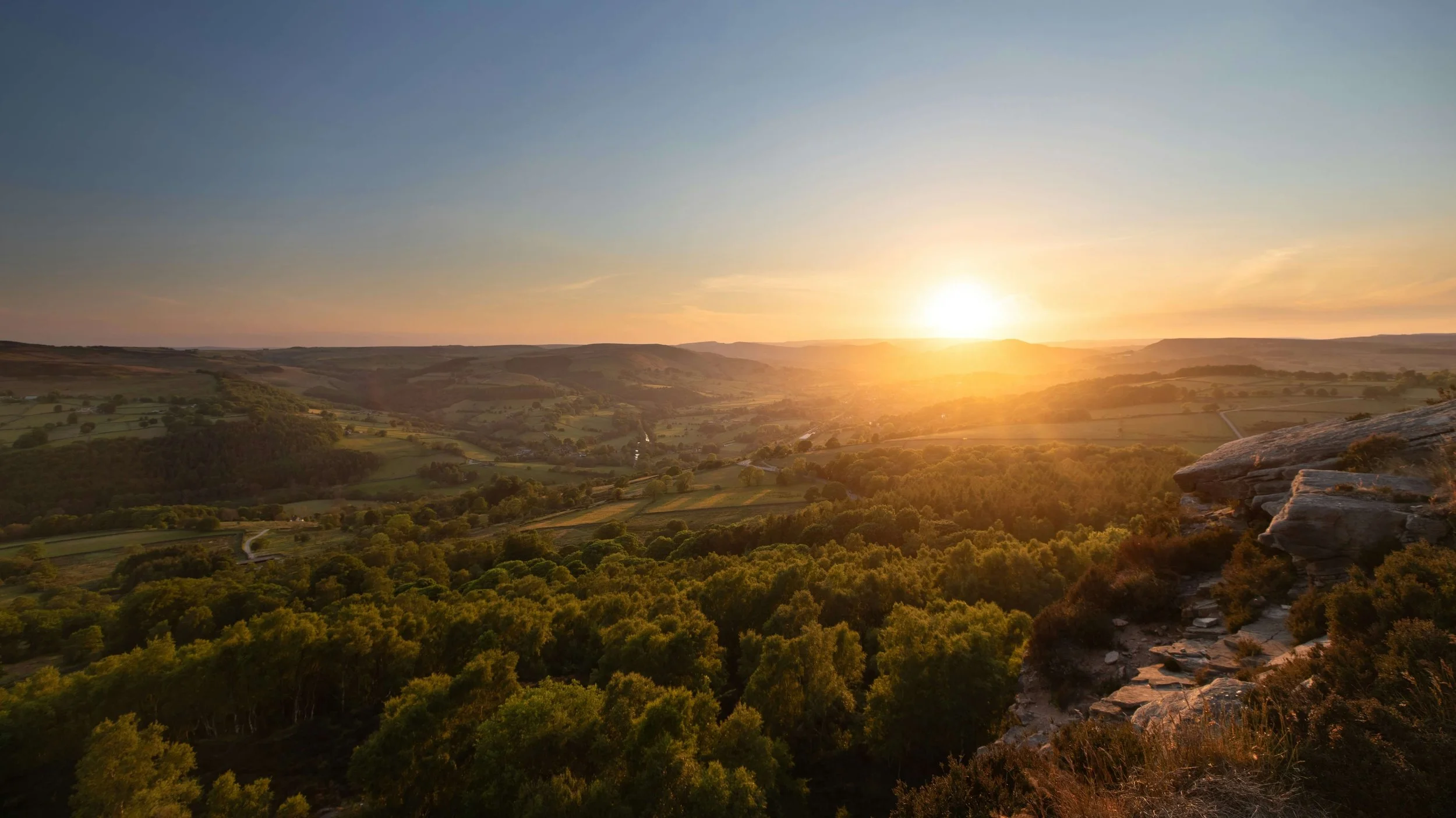 Sunset over rolling hills and lush green trees, with rocky outcrop on the right