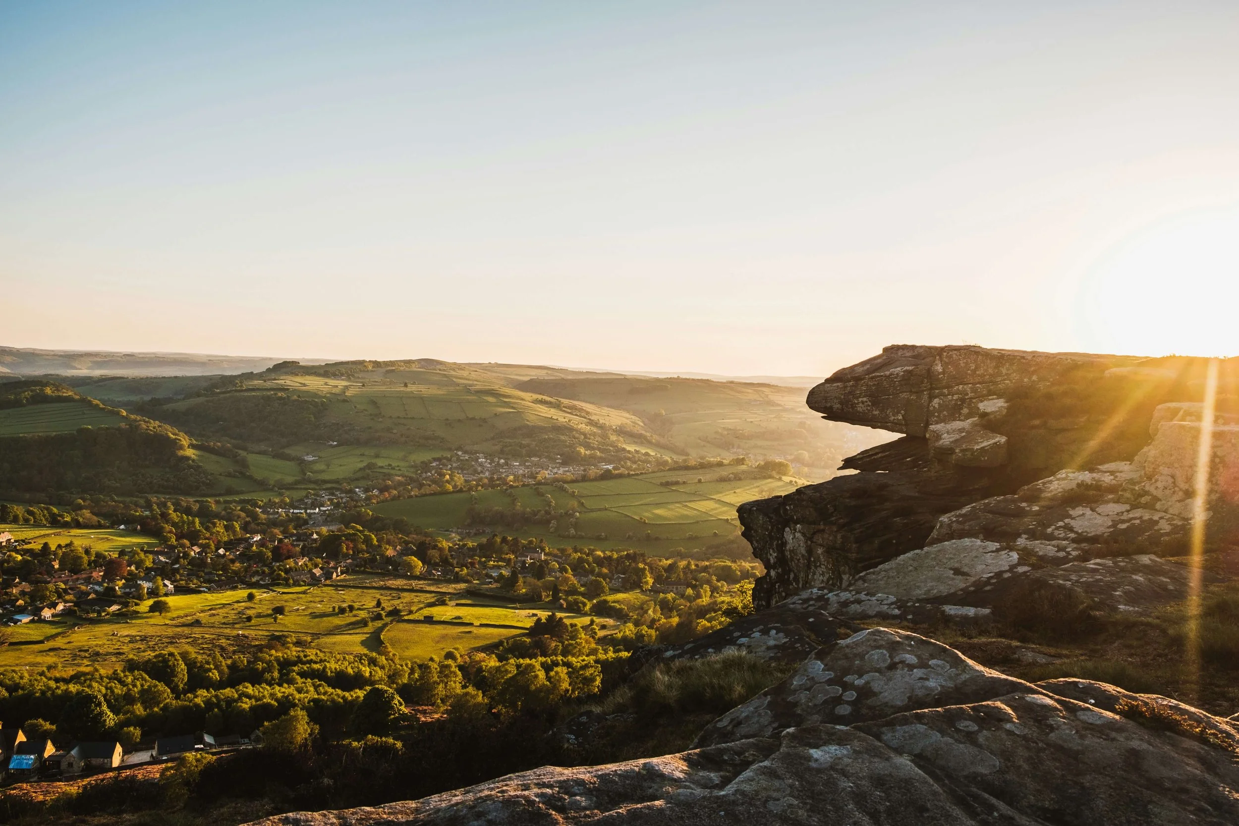 Sunset over a lush, green valley with rolling hills, trees, and a small village, viewed from a rocky cliff.