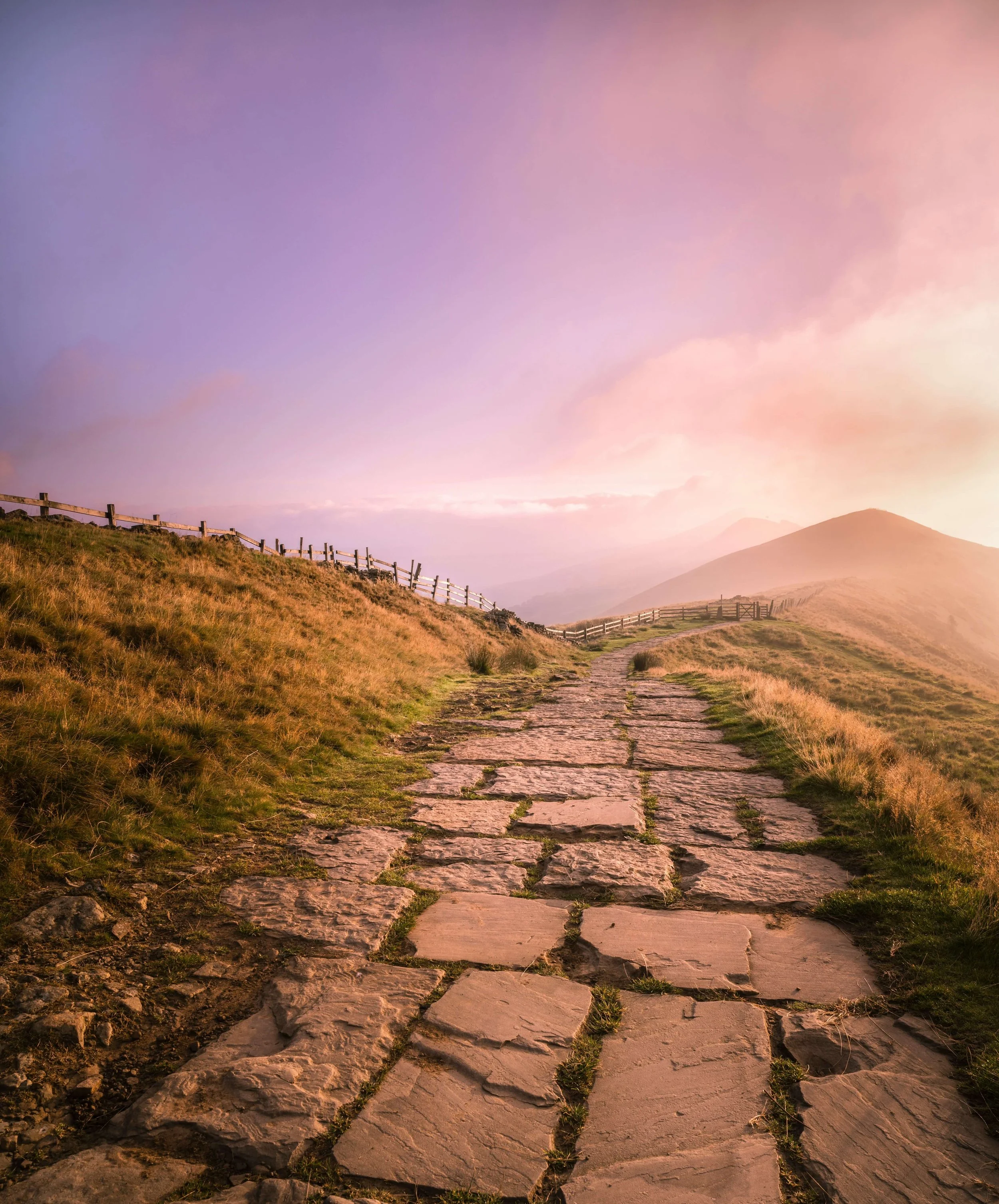 A stone pathway winding through a hillside with grassy terrain, wooden fences on the sides, and mountains in the distance under a pink and purple sky.