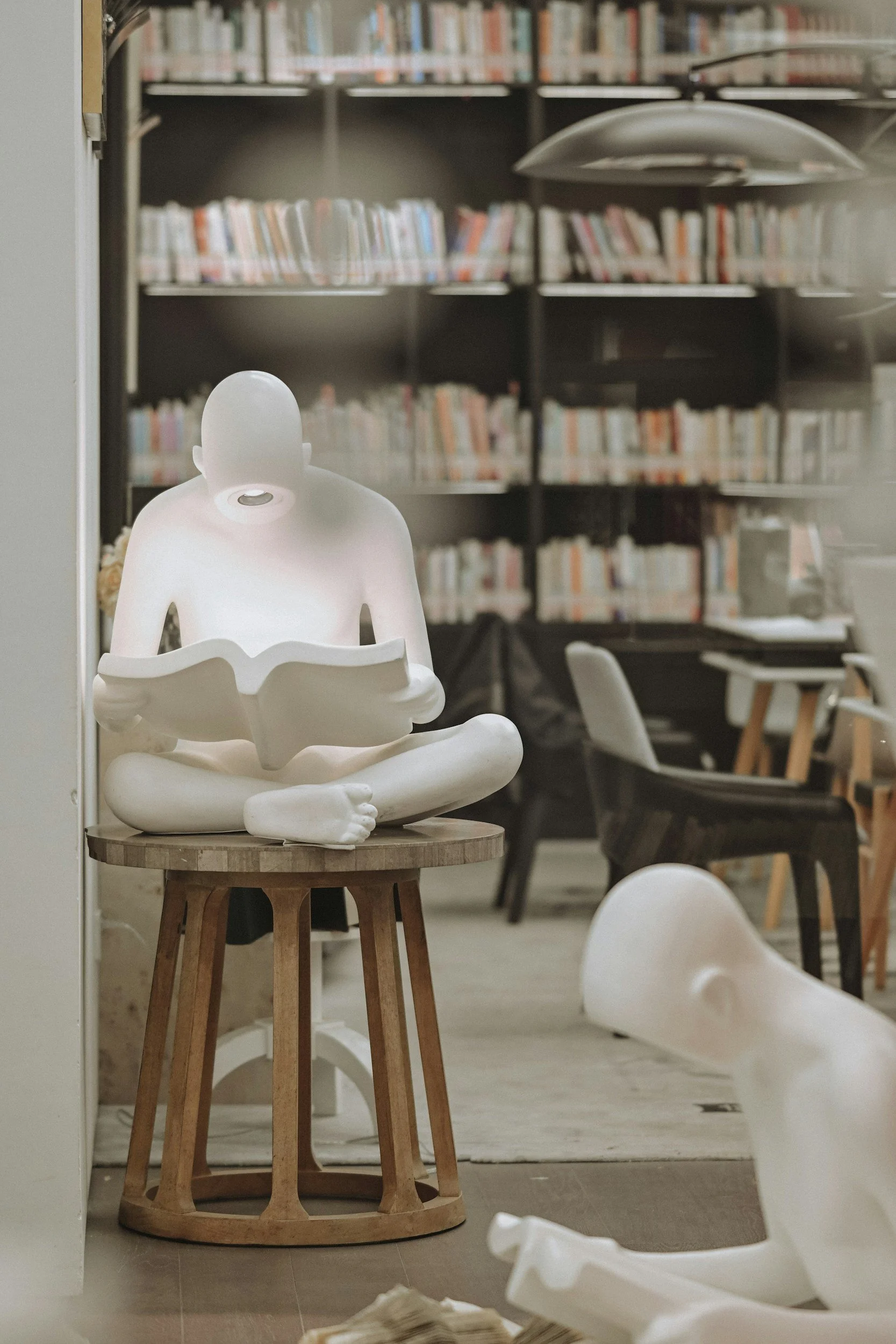 A white, sculpture-like figure of a person reading a book, sitting cross-legged, placed on a small round wooden table in a library or bookstore with bookshelves in the background. The scene includes white chairs and a black table, with a modern, minimalistic aesthetic.
