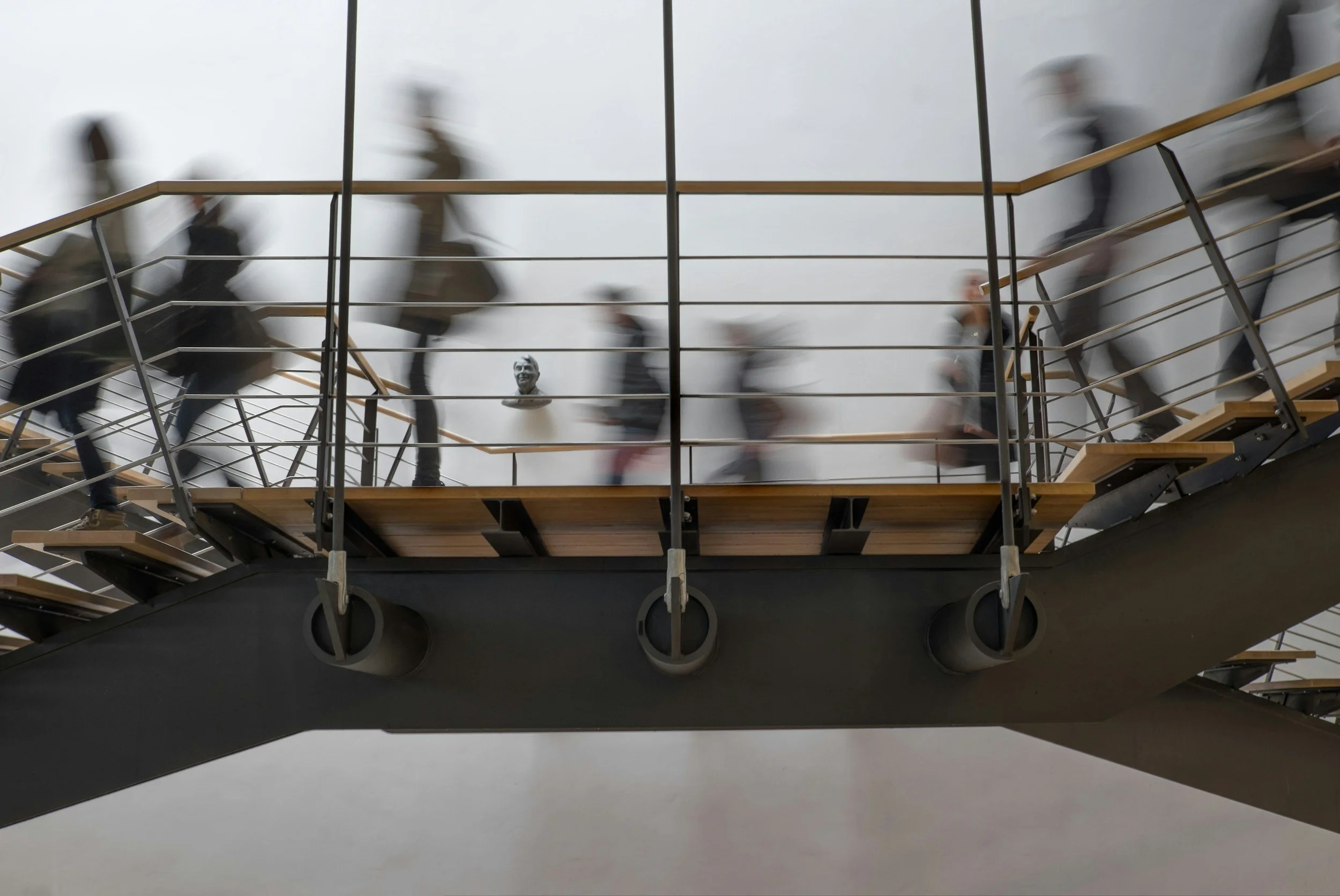 Blurred motion of people walking up and down a staircase with metal railings and wooden steps, inside a modern building.