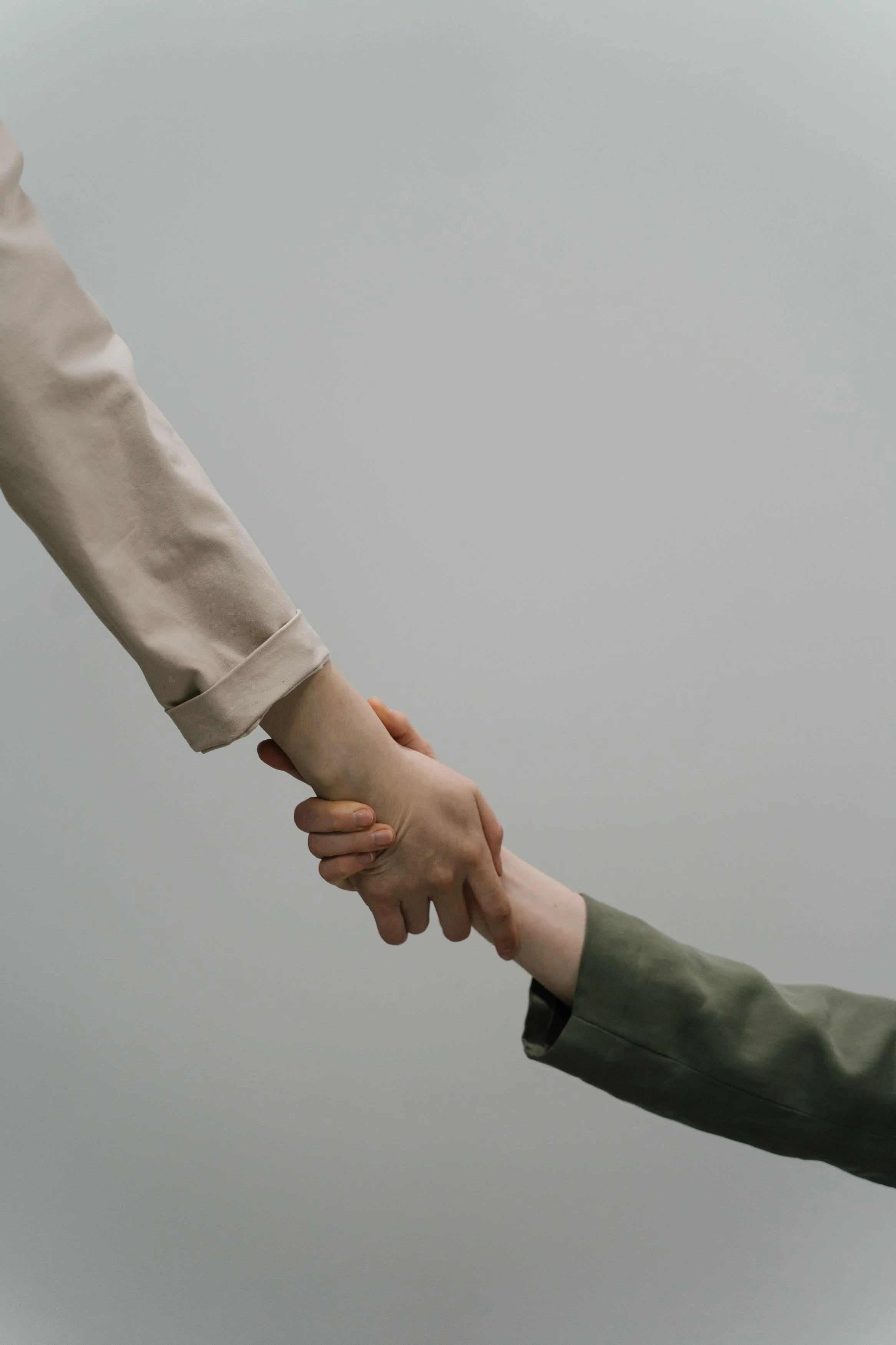 Two people holding hands, one wearing a beige jacket and the other wearing a grey jacket, against a plain light-colored background.