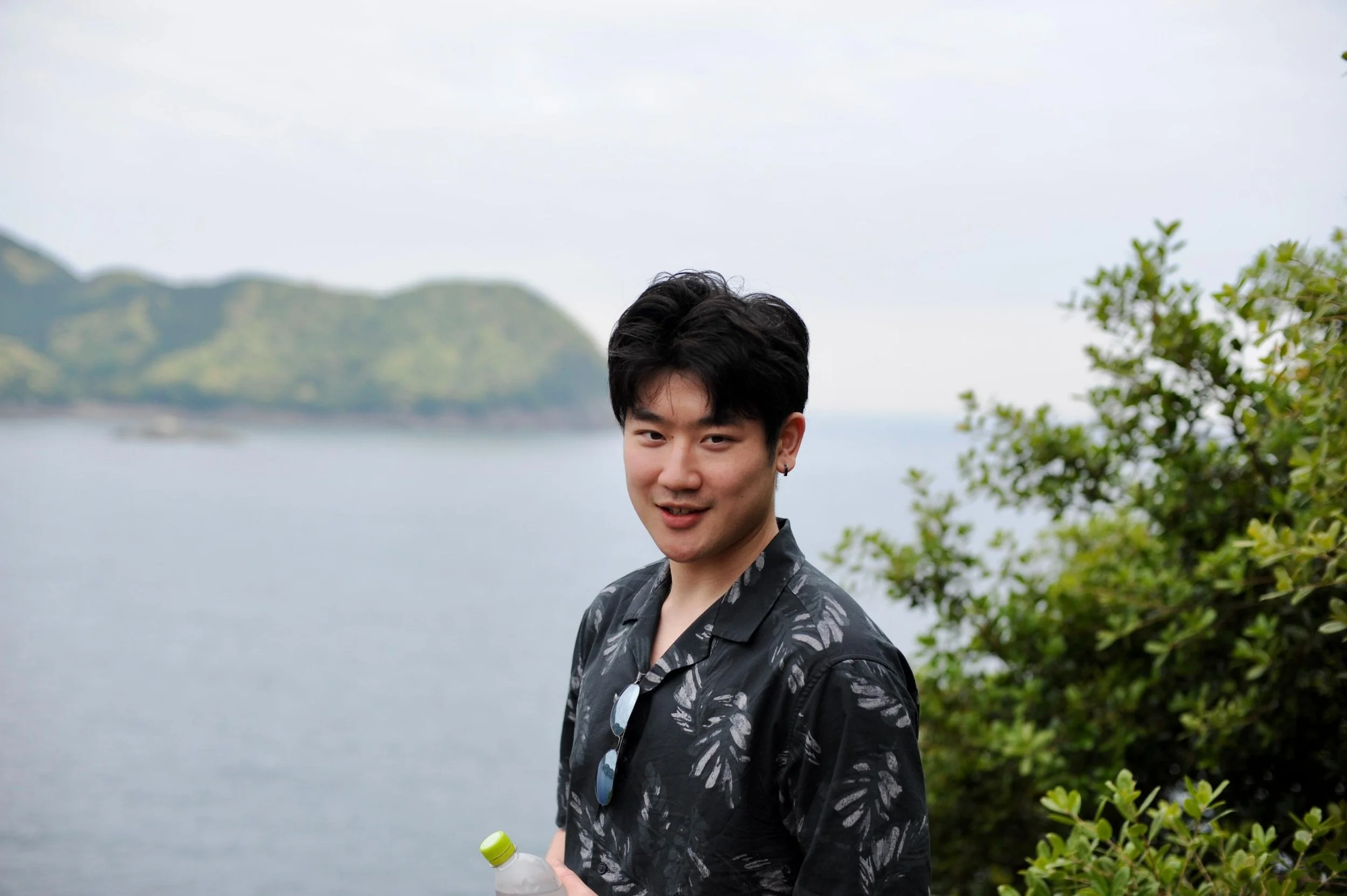 A young man with black hair standing outdoors near the water, holding a water bottle, with green foliage on the right and a mountain in the background.