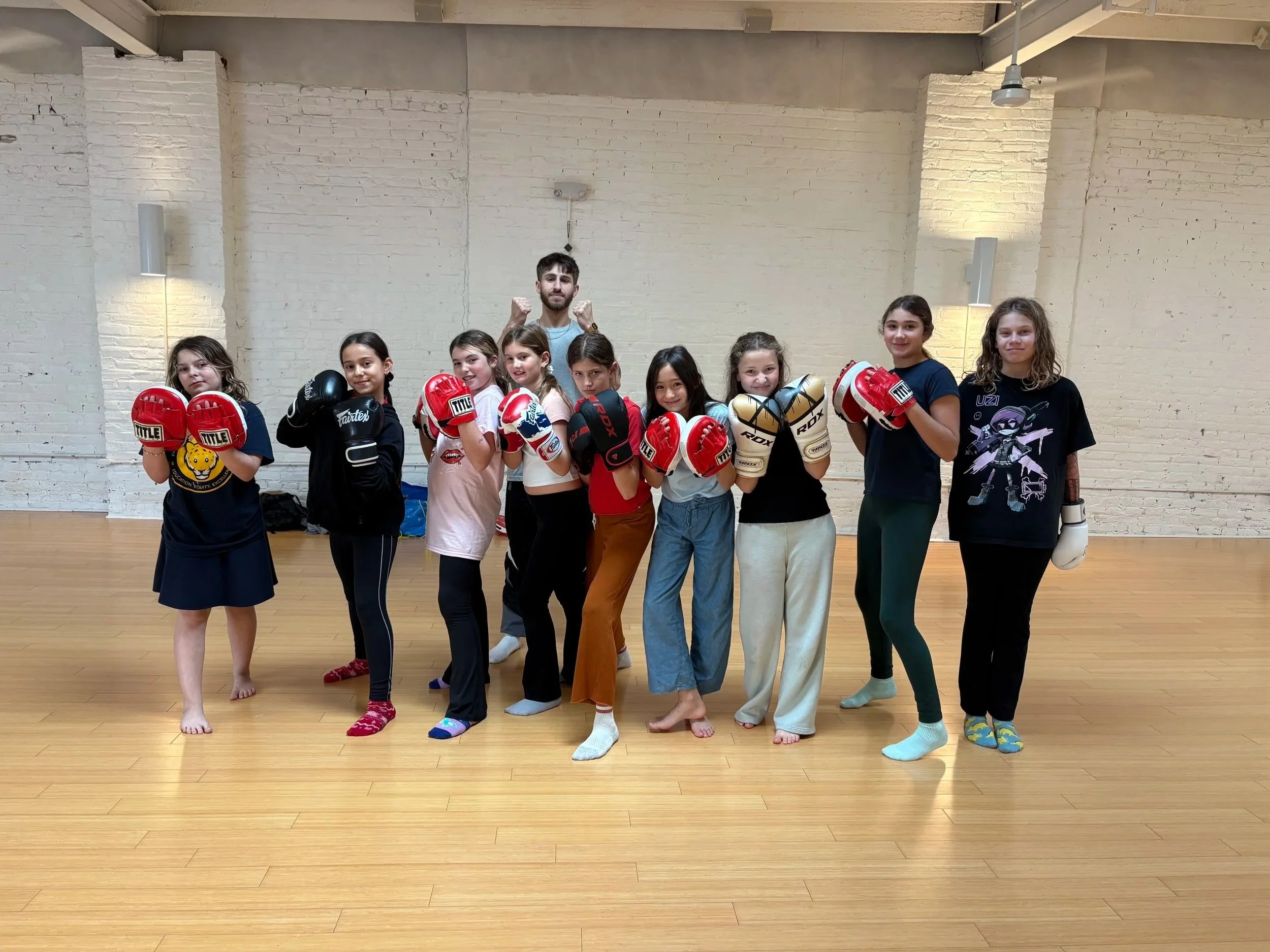 Group of girls and a male instructor in a martial arts studio, dressed in casual clothes and wearing boxing gloves, standing in fighting stances on a wooden floor with a white brick wall background.