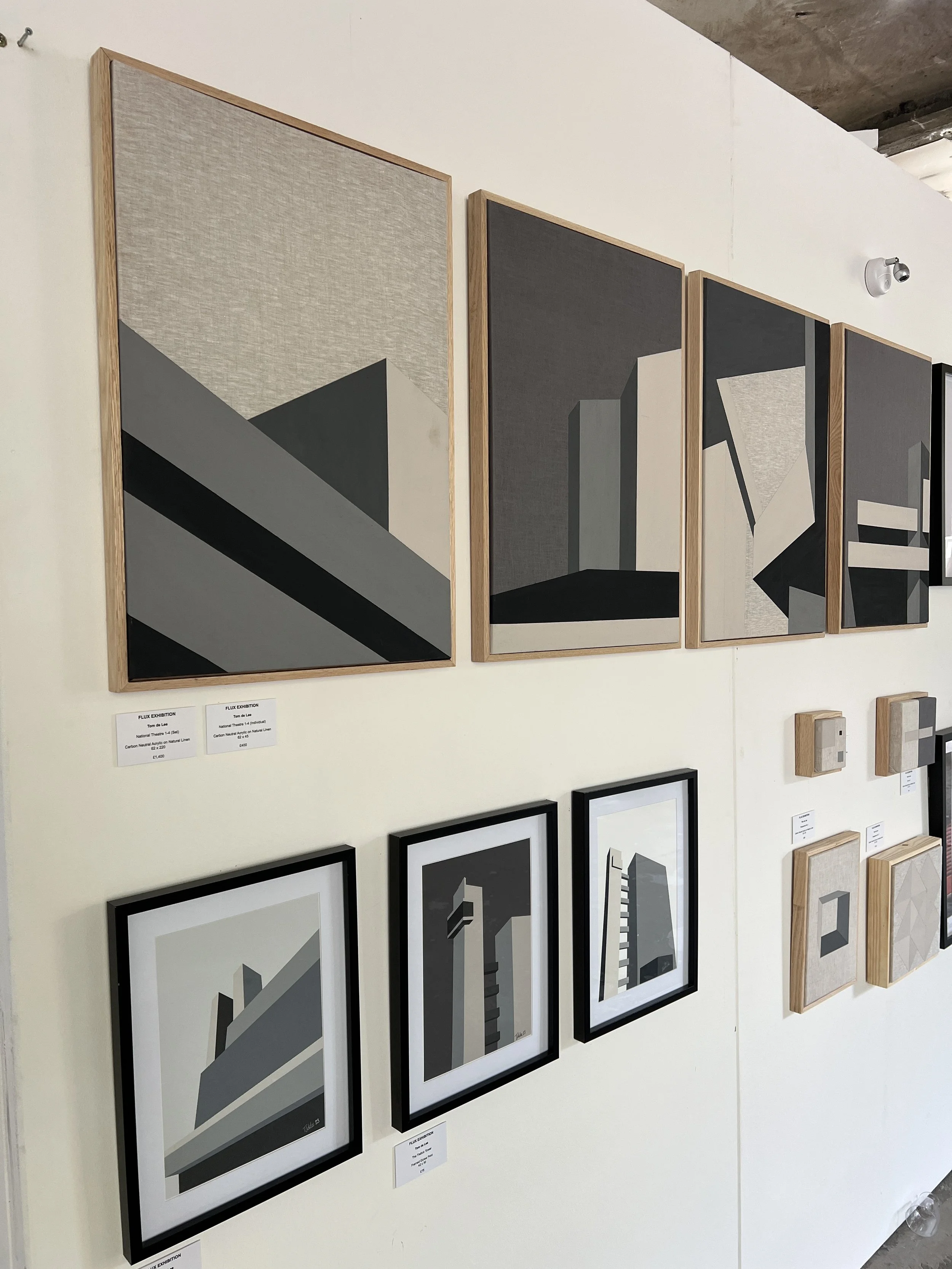Collection of modern, abstract artwork featuring geometric shapes in various shades of gray, black, and white displayed on a gallery wall.