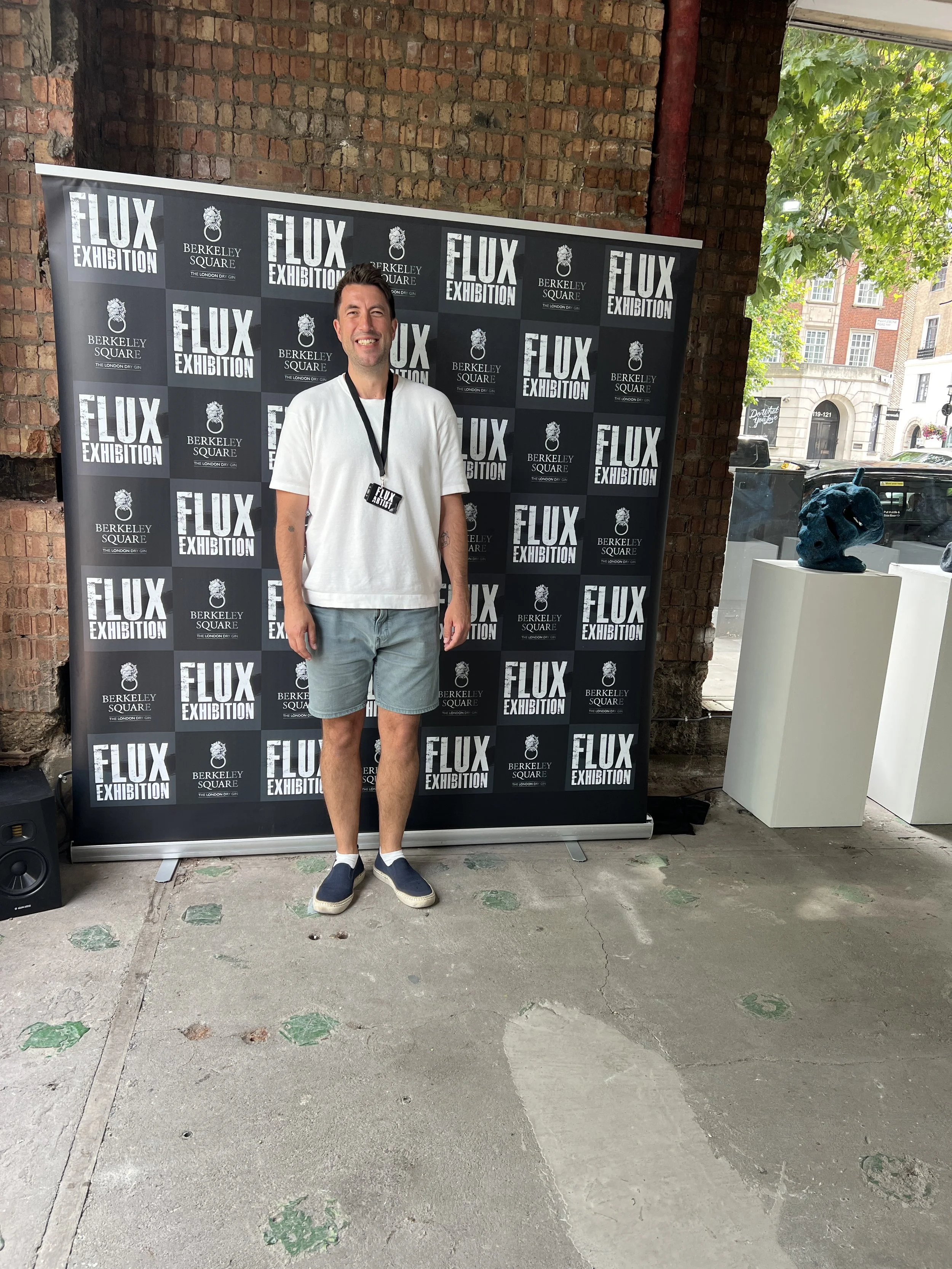 A man standing in front of a black and white checkered backdrop with the words 'Flux Exhibition' and 'Berkeley Square' written on it. He is smiling and wearing a white t-shirt, gray shorts, and navy sneakers, with a lanyard around his neck. To the right, there is a white pedestal displaying a blue sculpture of a head. The background includes a brick wall and a window showing green trees outside.