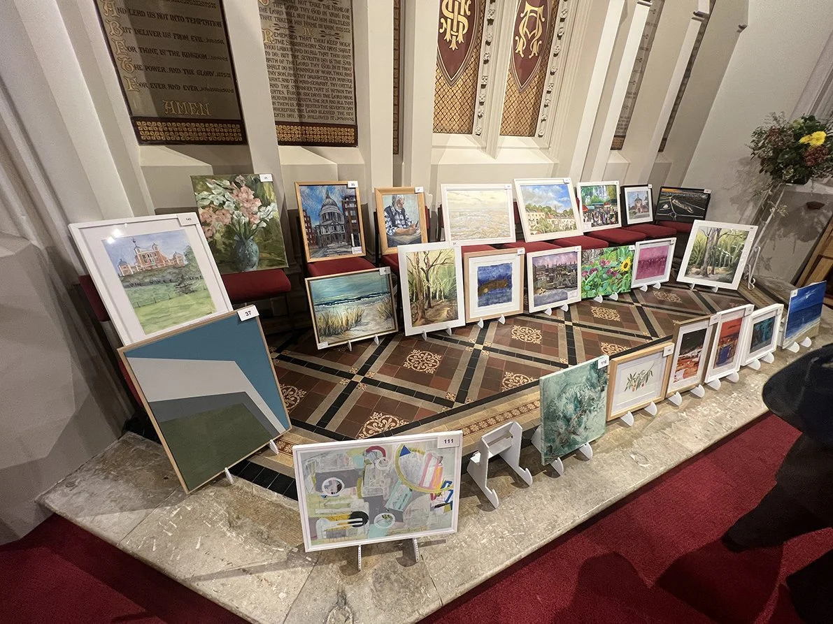 Display of framed paintings arranged on a tiered stand and table in an art exhibition space. The paintings depict various landscapes, cityscapes, floral scenes, and abstract art, set against the backdrop of an ornate interior with decorative banners and floral arrangements.