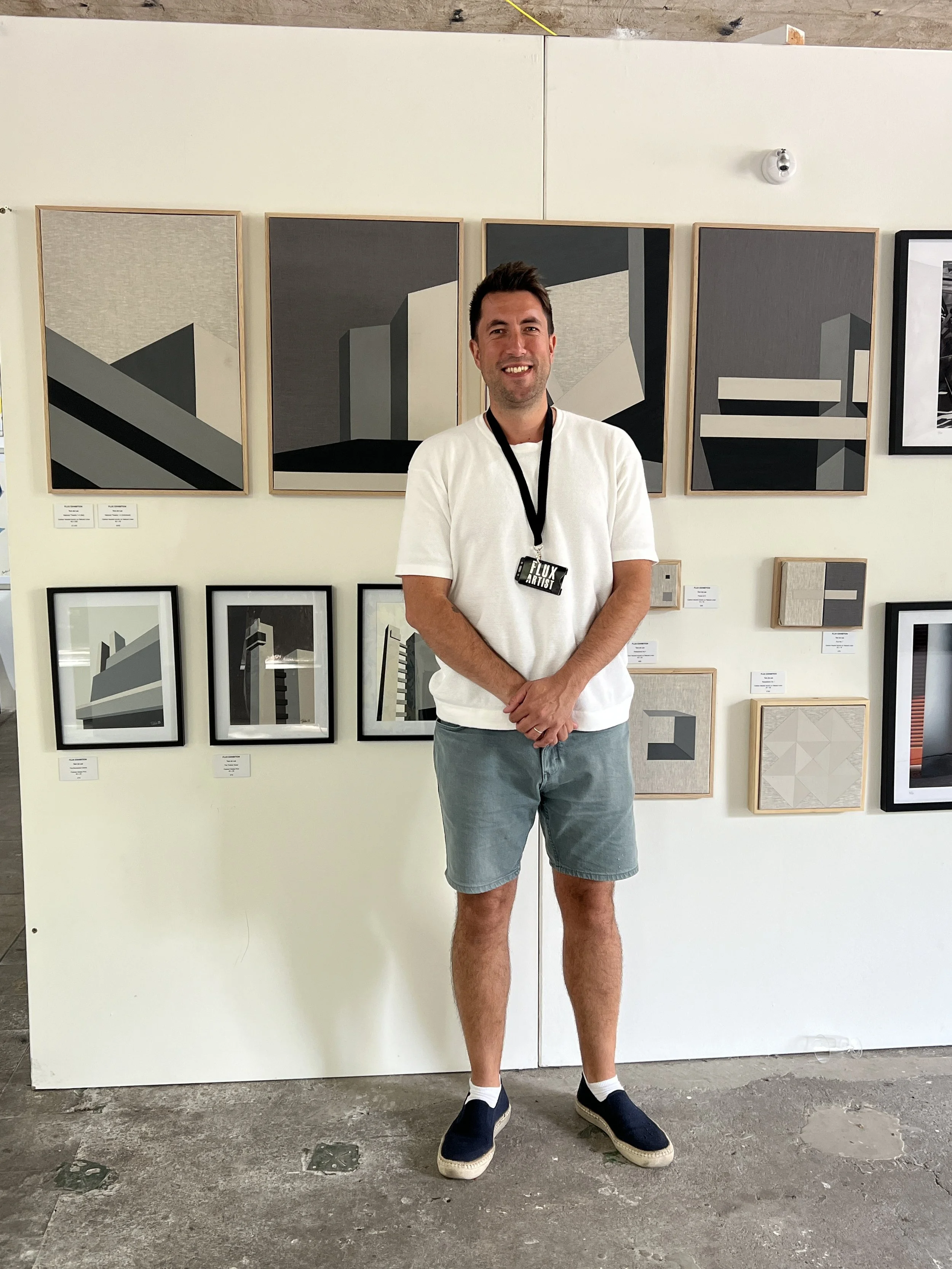 A man with short dark hair and light skin stands smiling in front of a wall with modern abstract artwork. He is wearing a white t-shirt, light blue shorts, navy slip-on shoes, and a lanyard with a badge that says 'Flux Artist'.