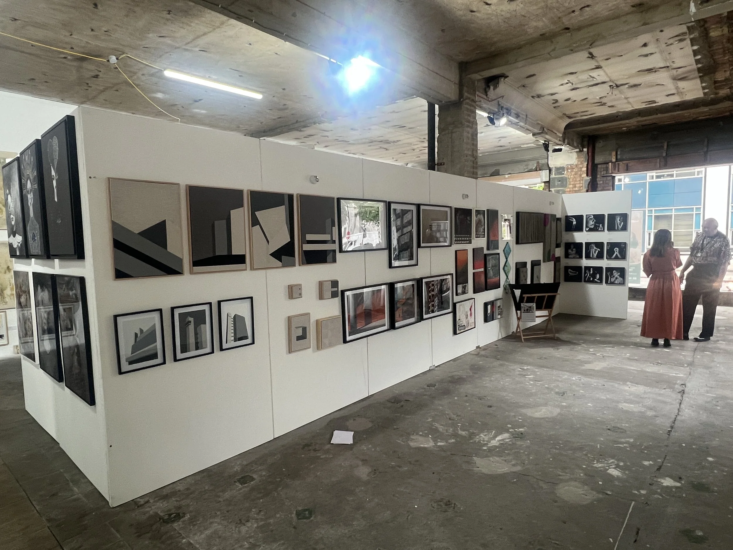 Art gallery with framed artworks on white walls, two people talking, exposed brick and industrial ceiling in the background.