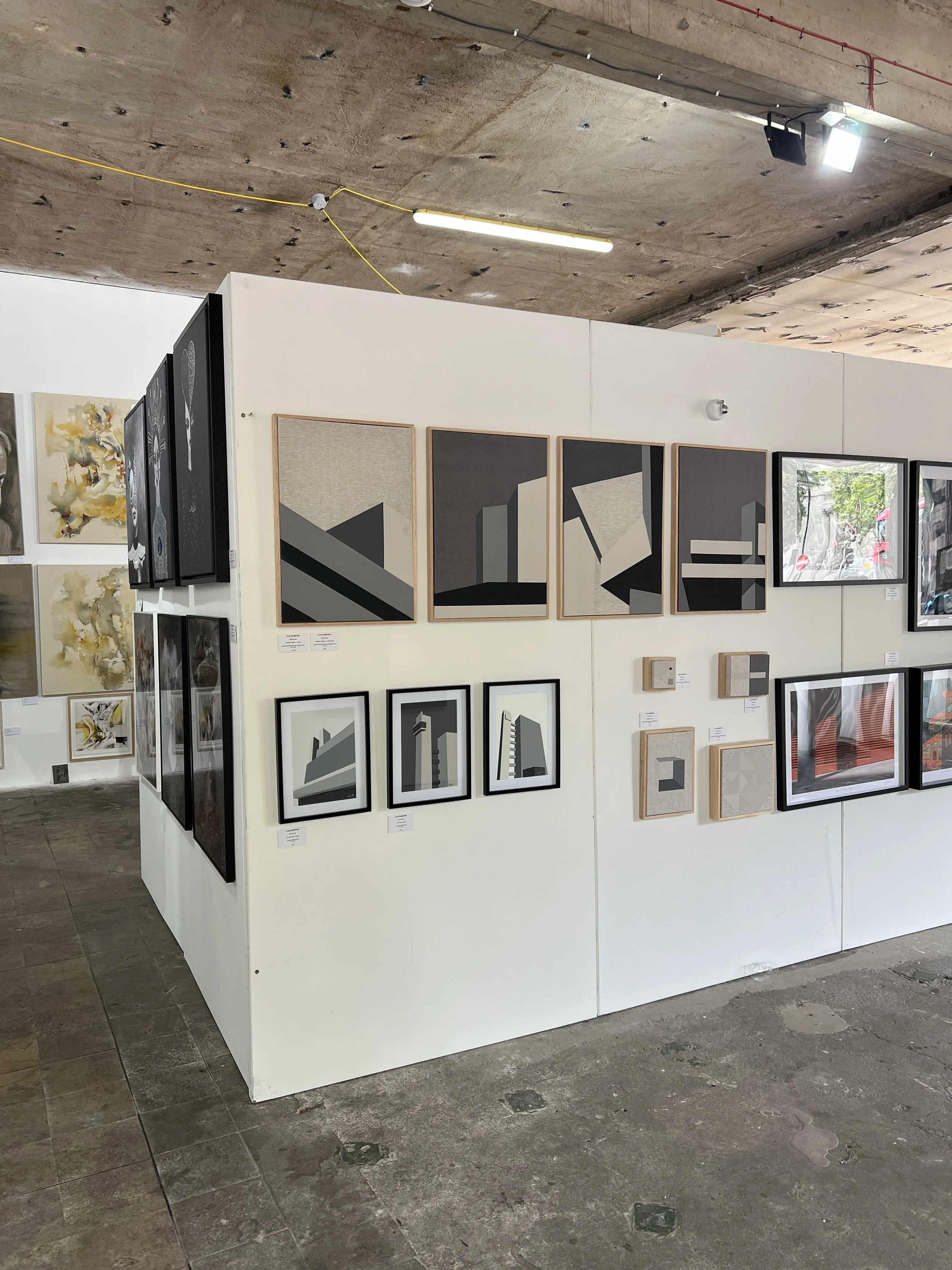 Art gallery display with various framed abstract and modern artworks on a white partition wall, including geometric shapes and muted color palettes, in an industrial-style setting with concrete flooring and ceiling.