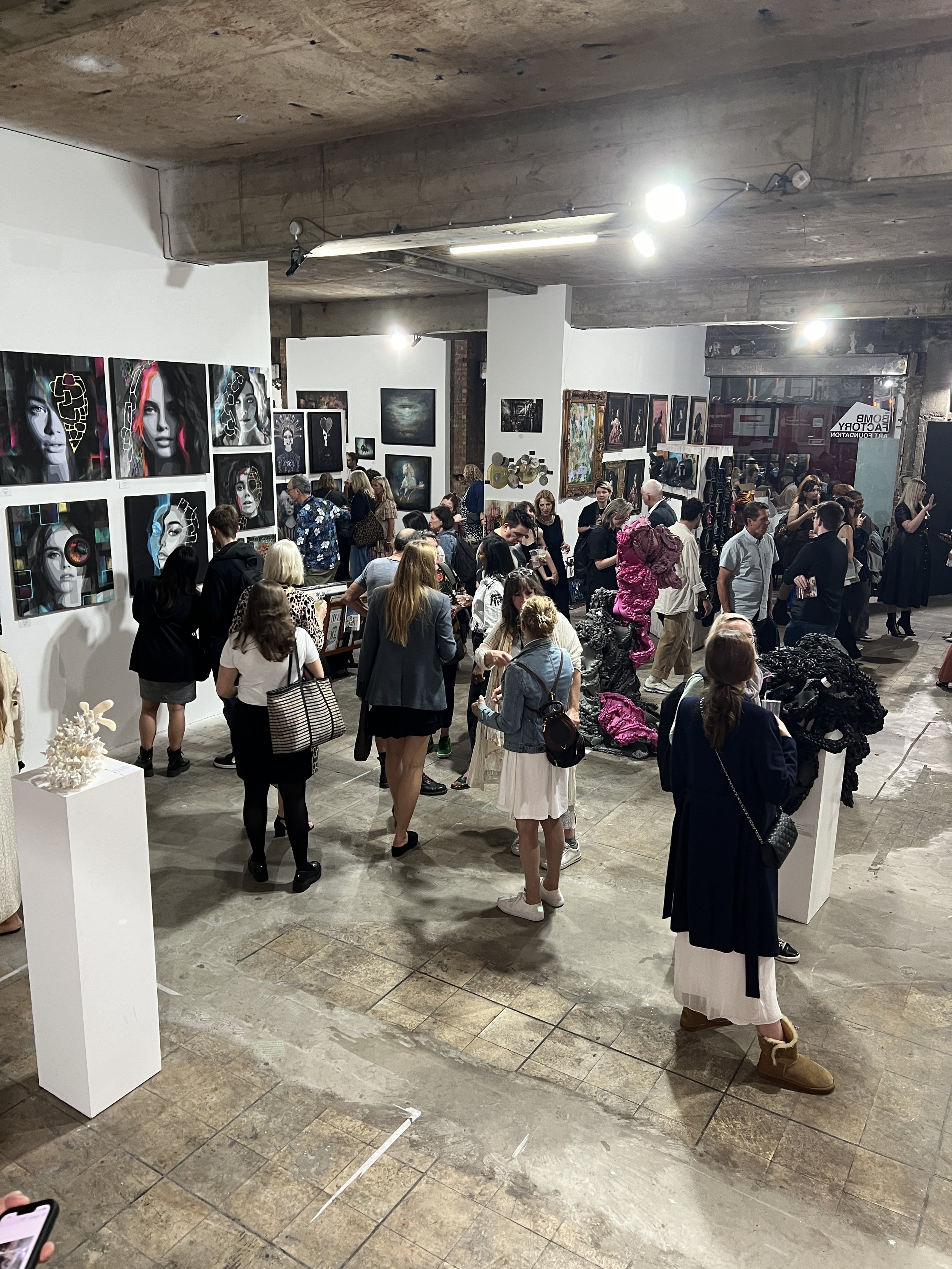 An art gallery or exhibition space filled with people viewing various artworks, including paintings displayed on white walls and sculptures on white pedestals. The gallery has an industrial look with exposed concrete floors and a wooden ceiling.