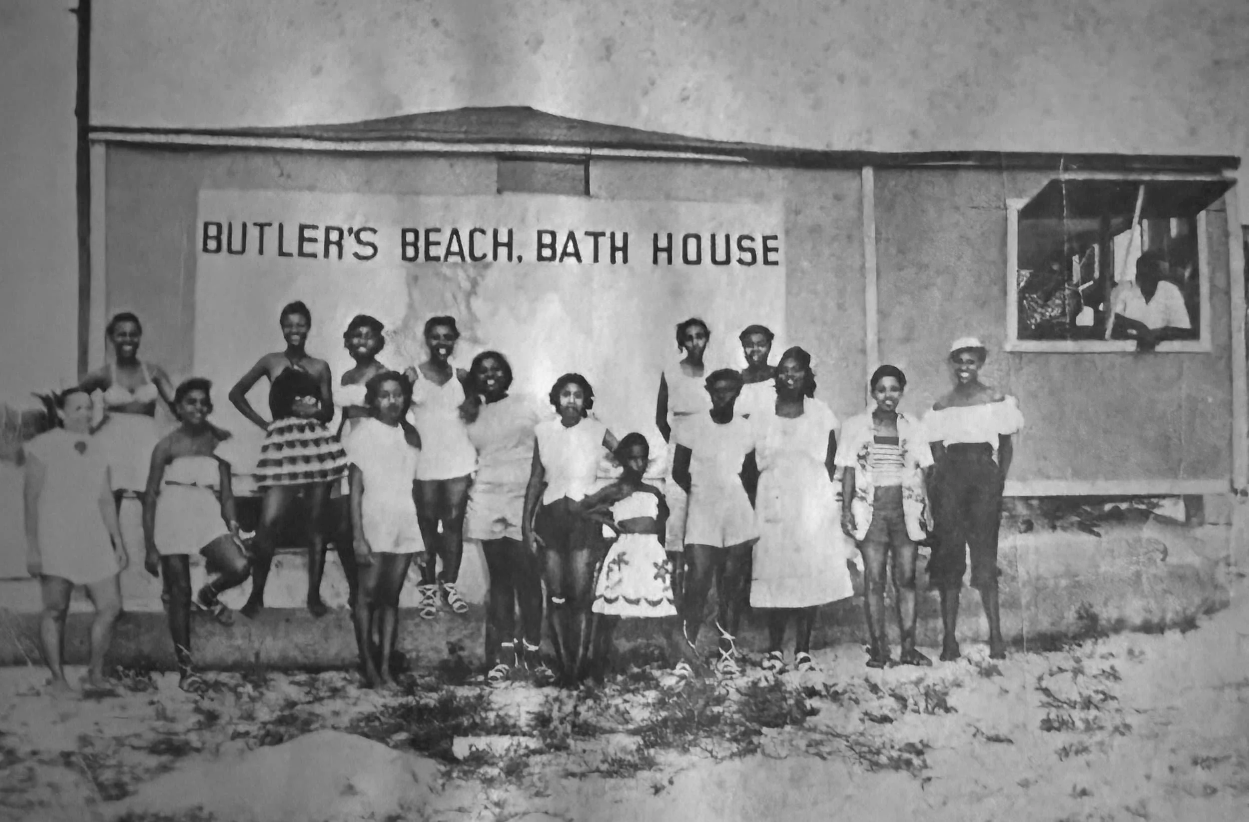 A group of people, including children and adults, standing in front of a building with a sign that reads 'Butler's Beach, Bath House.' The building appears to be a modest, single-story structure with a small window on the right, showing two people inside.