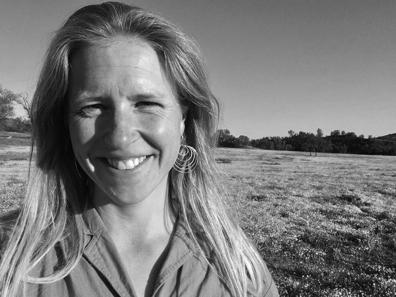 Black and white photo of a smiling woman with long hair, wearing earrings and a collared shirt, standing outdoors in a wide open field with trees in the distance.