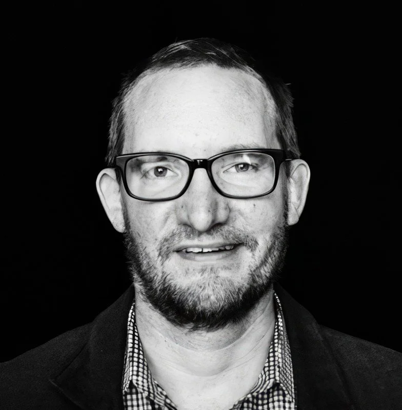 A black and white portrait of a man with glasses, short hair, and a beard, wearing a blazer and a checkered shirt.