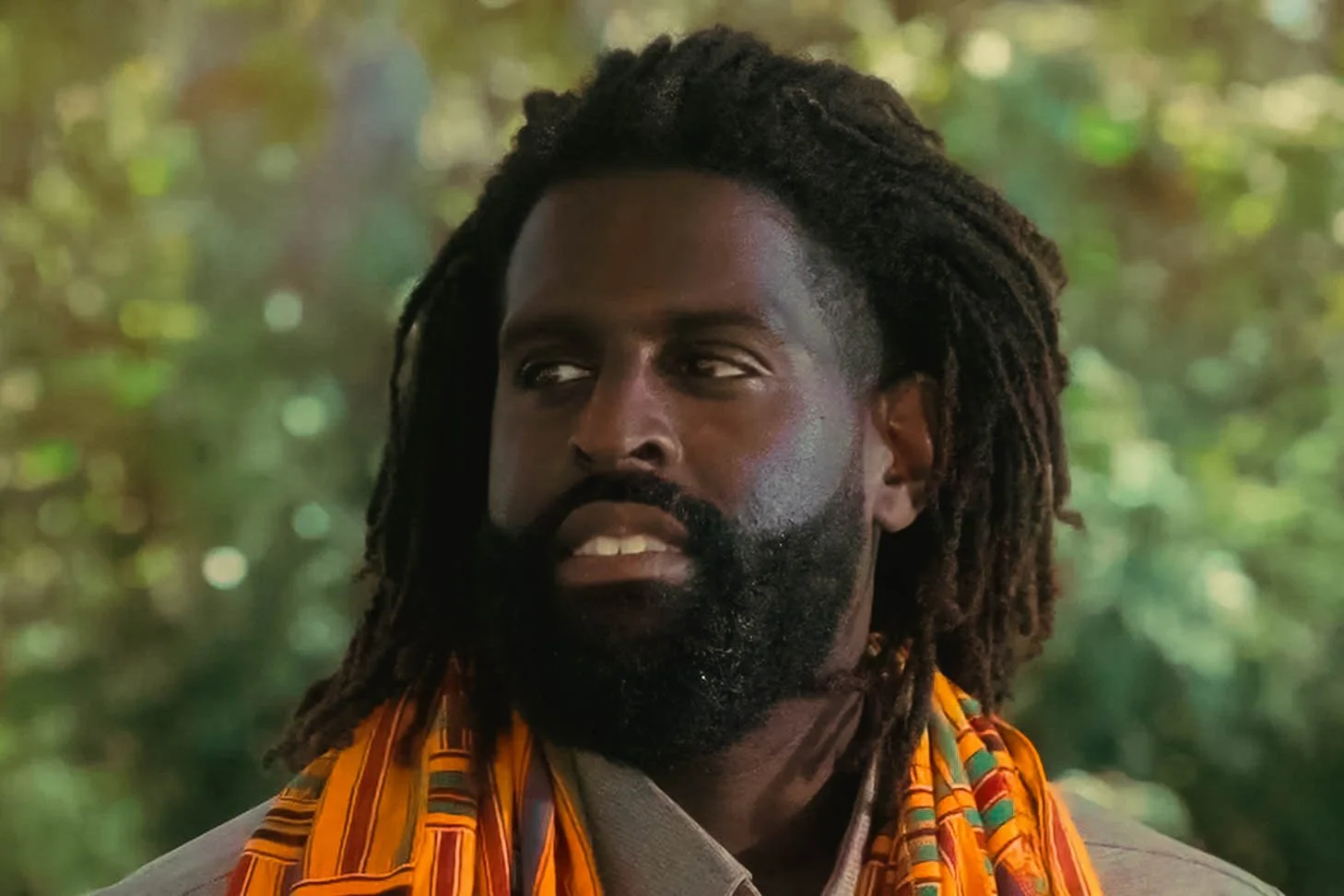 Close-up of a man with dark skin, dreadlocks, and a beard, wearing a colorful orange scarf, outdoors with blurred green foliage in the background.