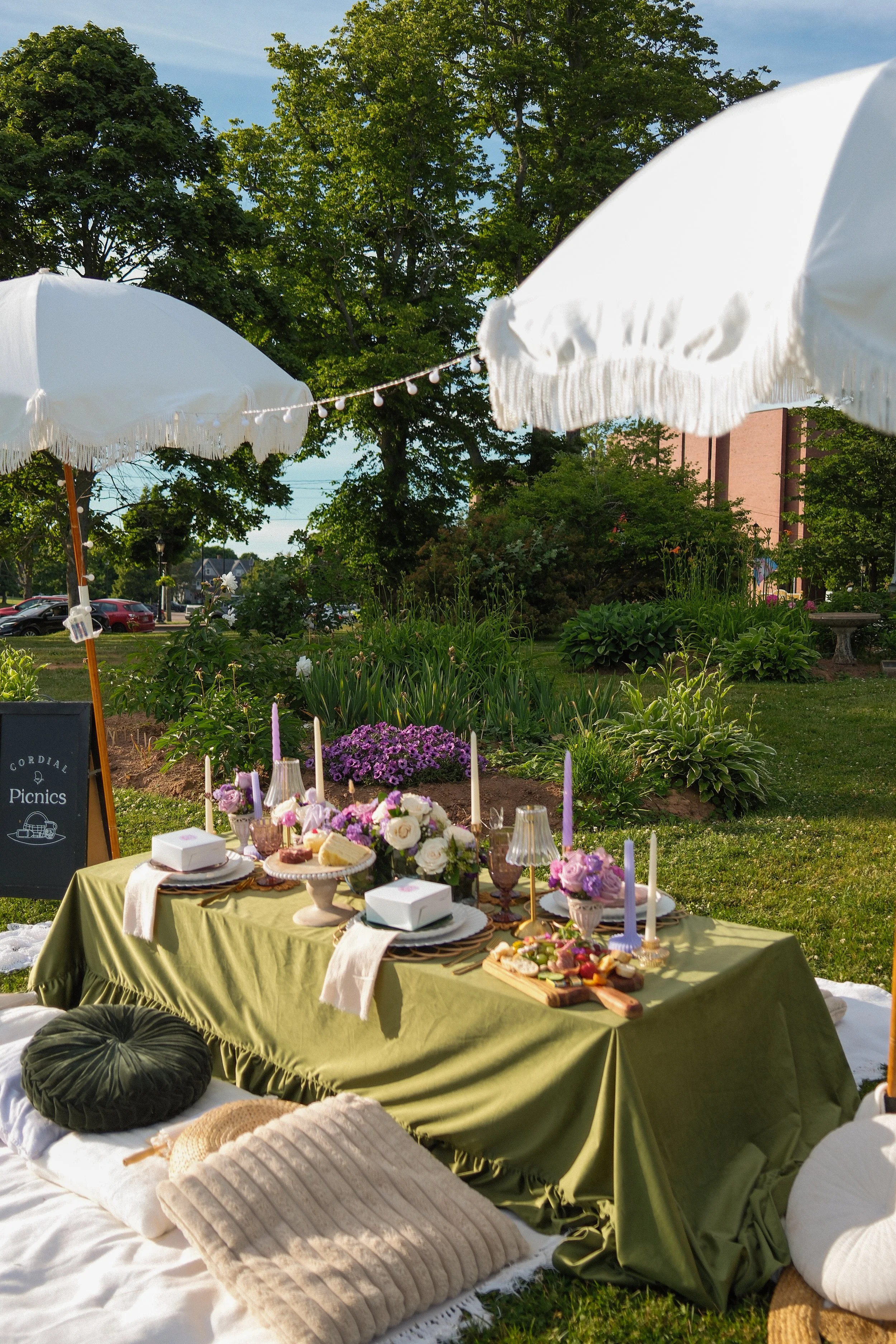 Outdoor picnic setup with a long table covered with a green tablecloth, decorated with flowers, candles, and picnic food, surrounded by cushions and white umbrellas, in a garden with trees and plants.