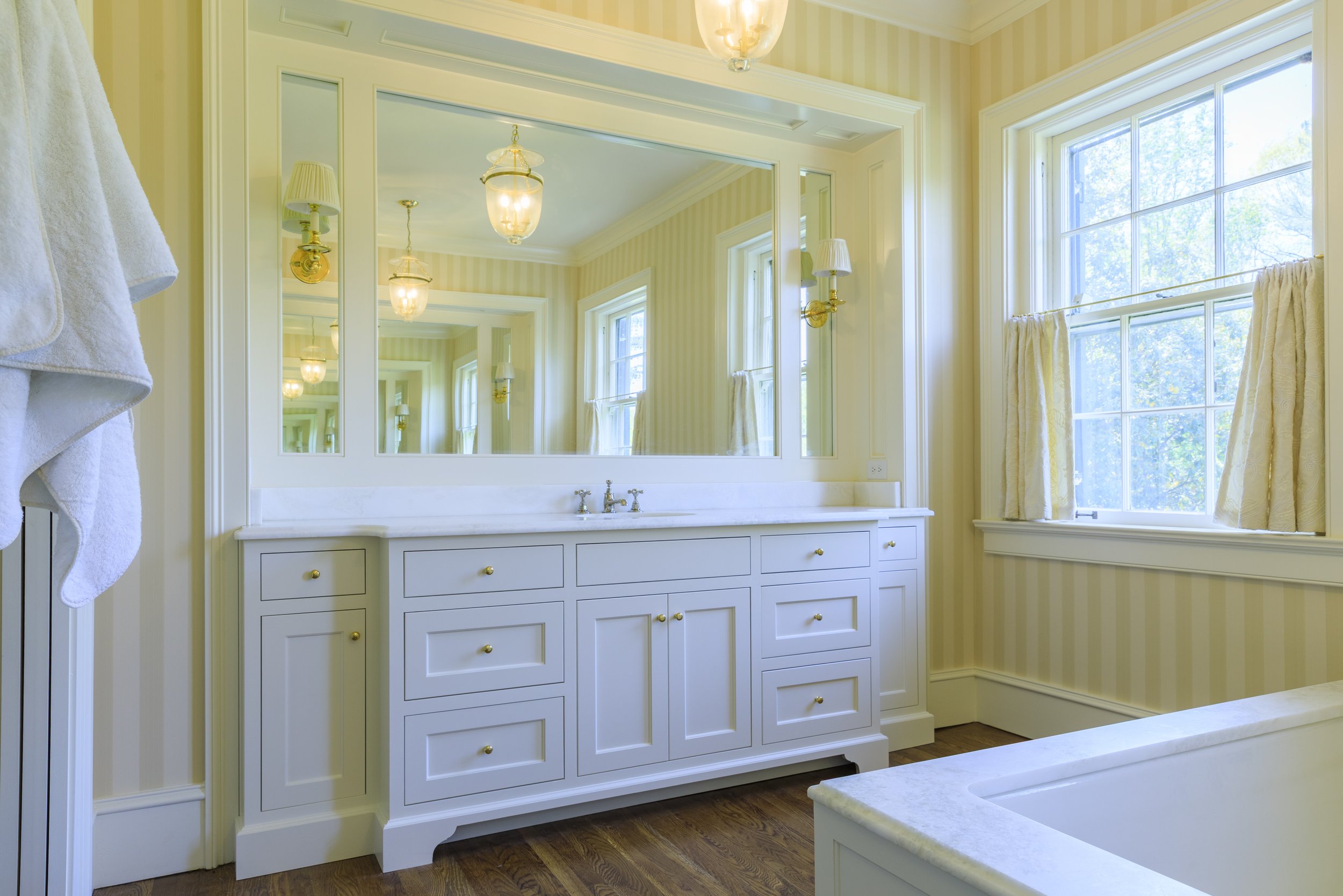Bright bathroom with a large mirror, white vanity with multiple drawers, a sink, and polished brass knobs. There are three windows with cream-colored curtains, and a wooden floor. The light fixture above has two glass lamps, and the walls are covered with light yellow striped wallpaper.