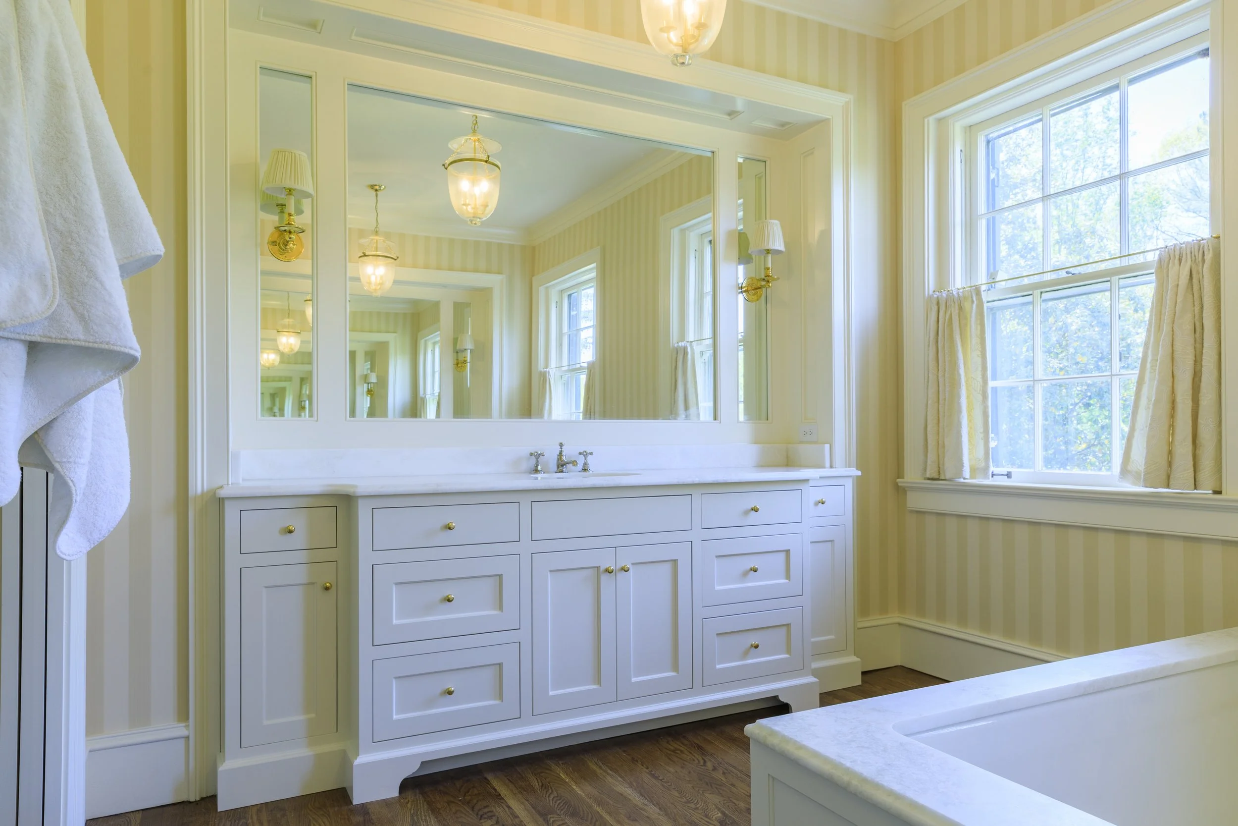 Bright bathroom with a large mirror, white vanity with brass knobs, and double sinks. Light yellow striped walls, hardwood floors, and a window with cream curtains letting in natural light.