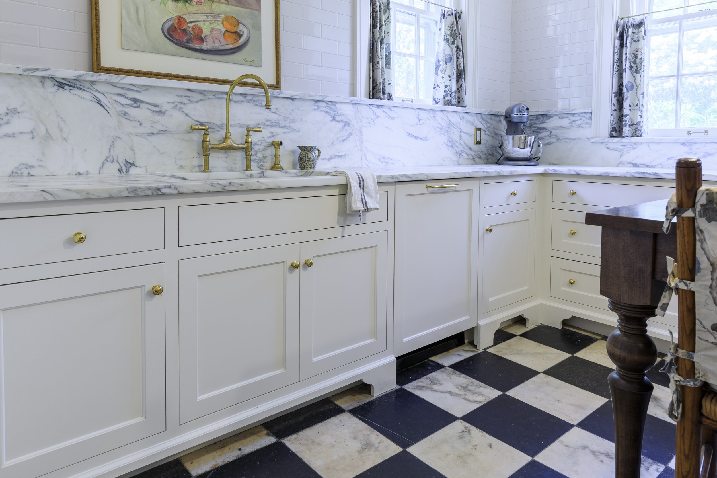 A kitchen with white cabinets and marble countertops, featuring a gold-colored faucet, a dish towel, and a stand mixer. There are two windows with floral curtains, and a framed still life painting of fruit on the wall. The floor has a black and white
