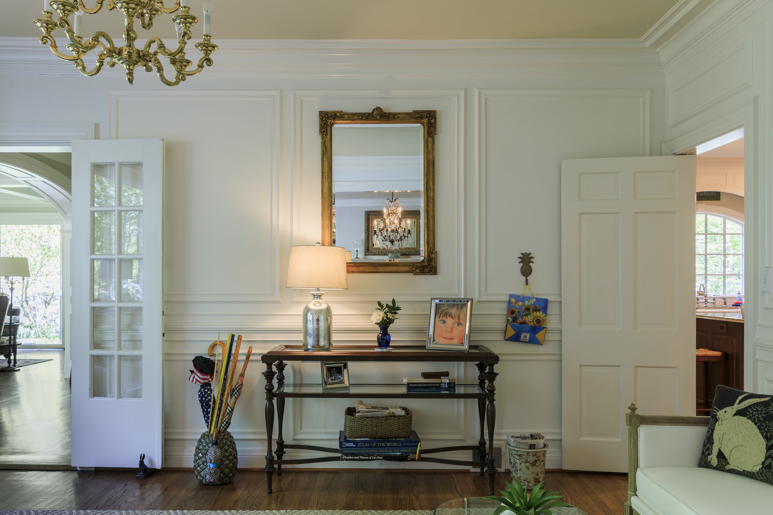 Entryway console table with lamp, framed photo, small plant, and books, mirror above, open door to living room and kitchen.