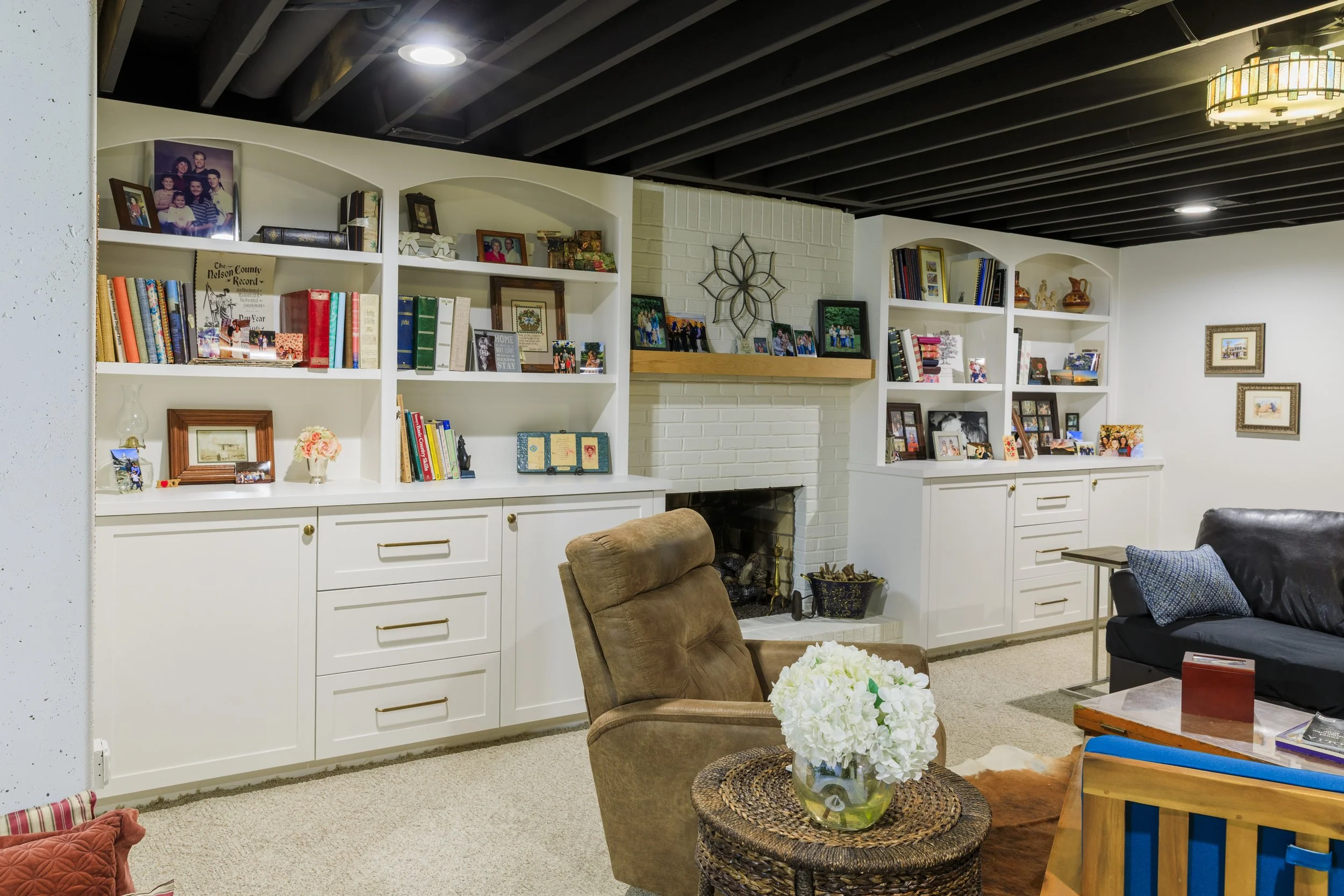 Living room with built-in white bookshelves filled with framed photos, books, and decorative items, a white brick fireplace with a wooden mantel, a tan armchair with a floral pillow, a black leather sofa with a blue pillow, a glass vase with white fl