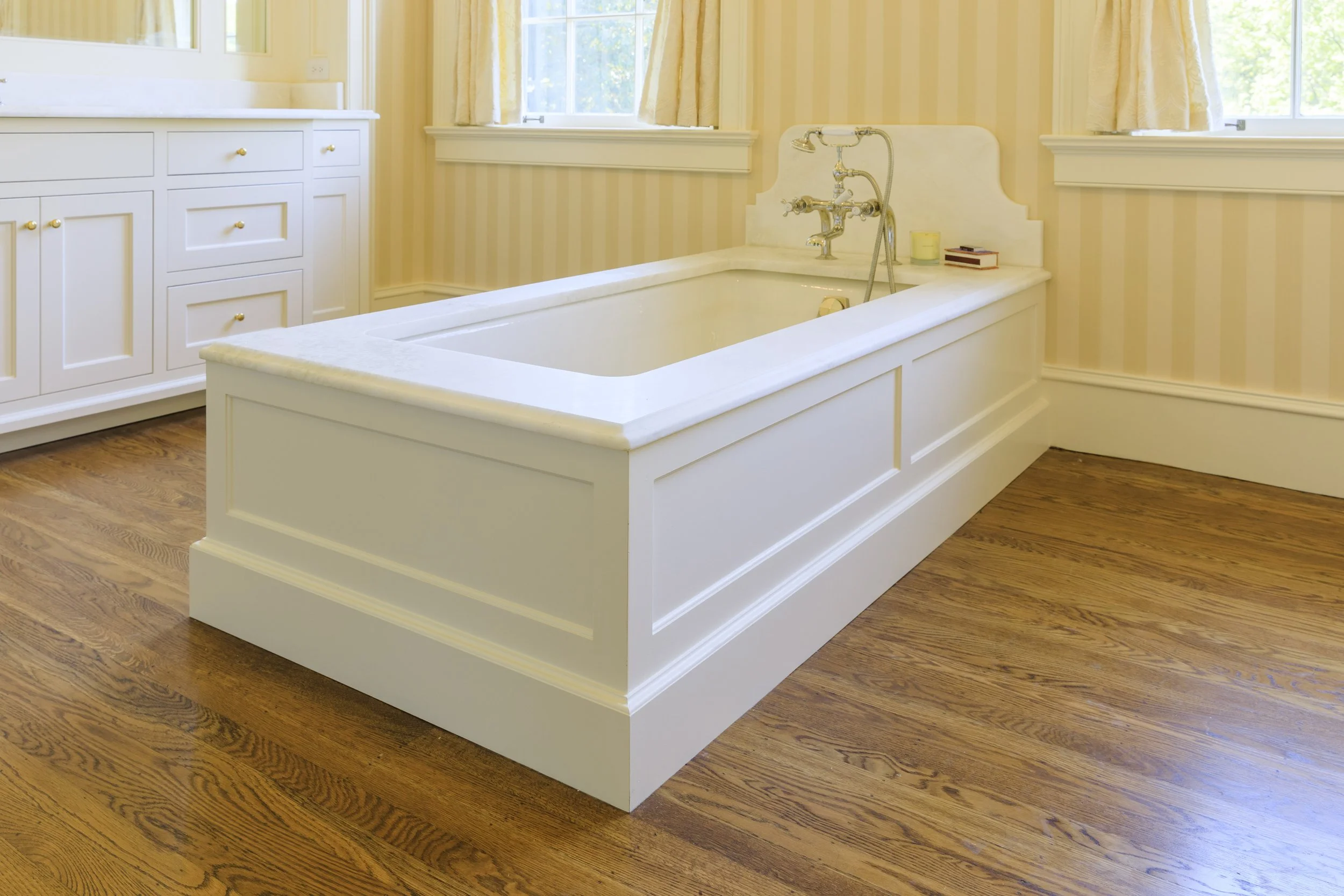 A white bathtub with a decorative wooden panel around it in a bright bathroom with hardwood floors and cream striped wallpaper, two windows, and built-in cabinets.