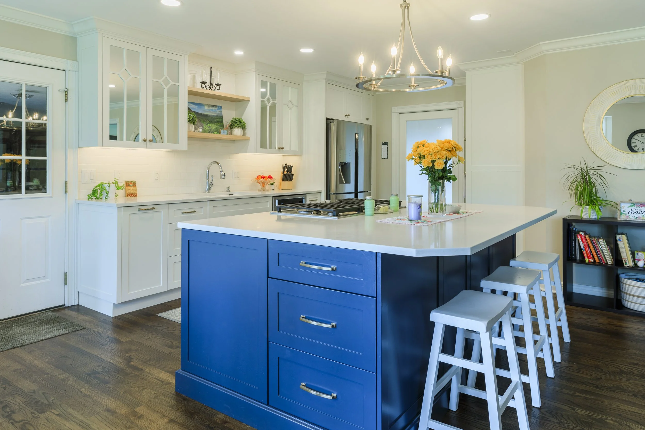 Modern kitchen with a large blue island, white countertops, and white cabinets, featuring a vase of yellow roses on the island and bar stools.