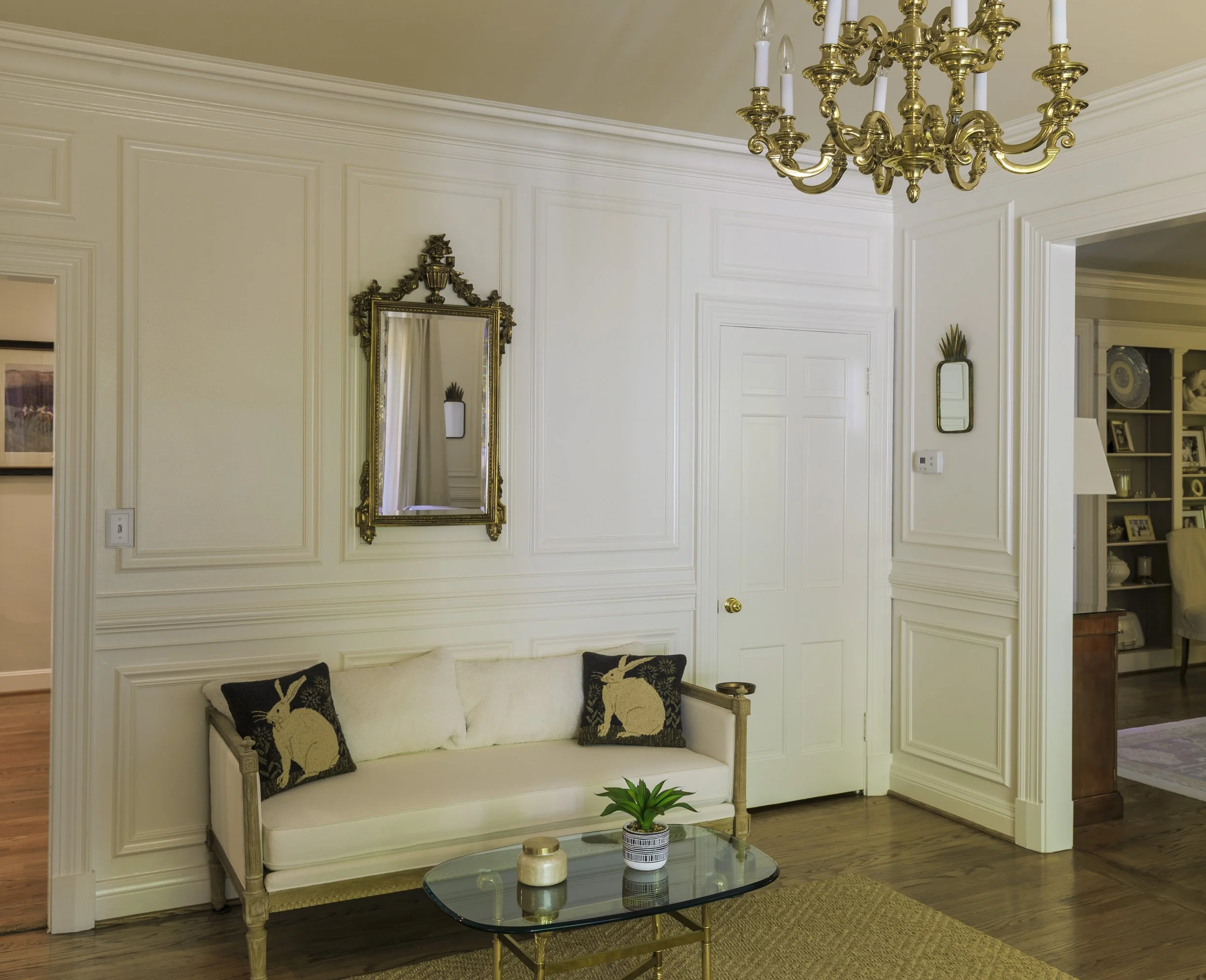 Elegant living room with cream-colored walls, gold chandelier, mirror with ornate frame, small sofa with bunny-themed throw pillows, glass coffee table with decorative items, adjacent to a doorway leading to a room with built-in shelves showcase deco
