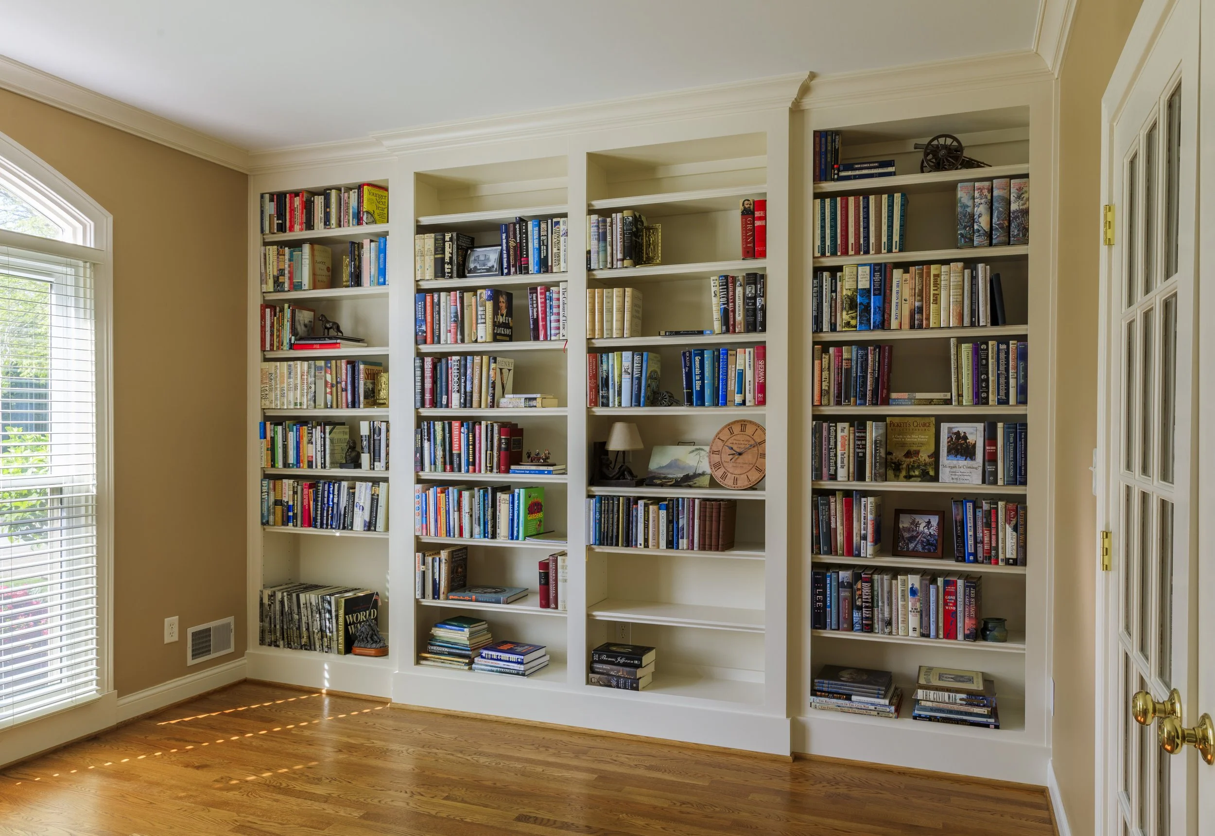 White built-in bookshelves filled with books, a small lamp, a clock, and decorative items in a room with hardwood floors and a large window with blinds.