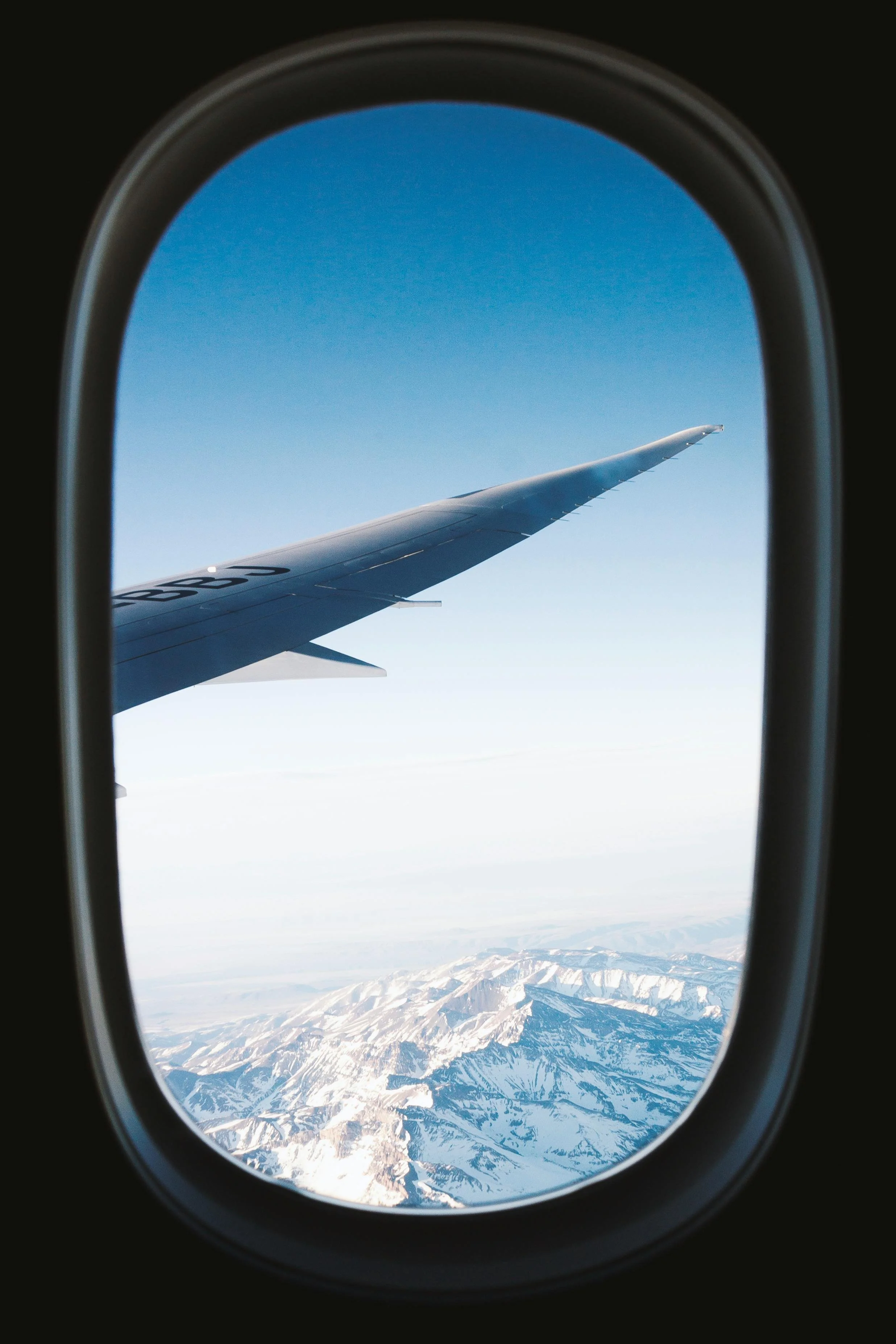 View of snowy mountains seen through airplane window with part of airplane wing visible.