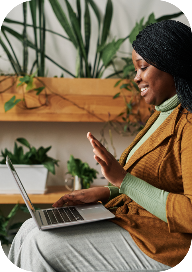 A woman smiling and waving at her laptop in a cozy indoor setting with green plants.