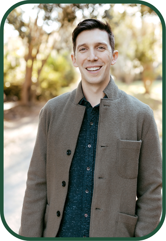 A smiling young man standing outdoors in a park, wearing a gray coat over a dark button-up shirt, with trees and sunlight in the background.