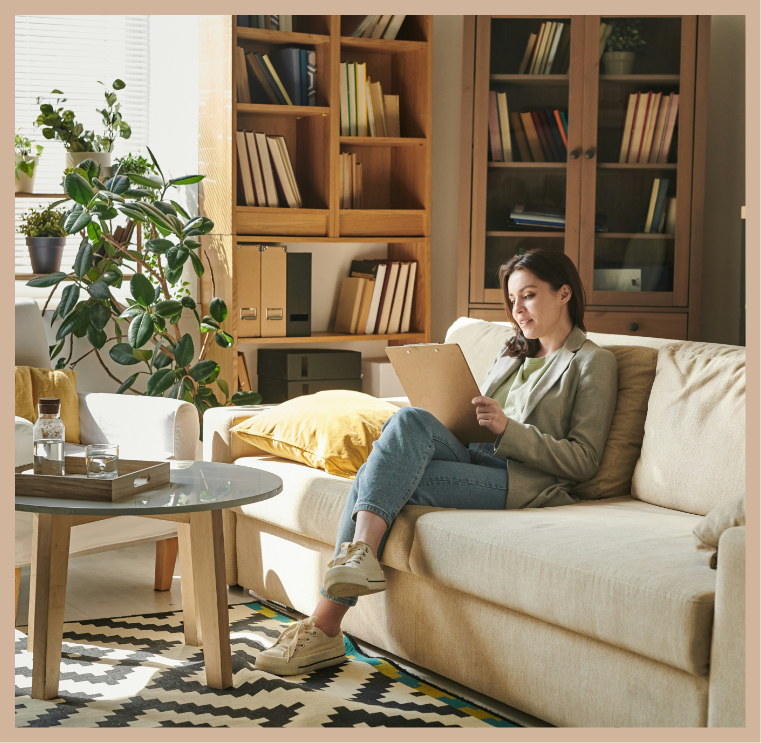 A woman sitting on a beige couch reading a clipboard in a cozy living room filled with books, plants, and sunlight.