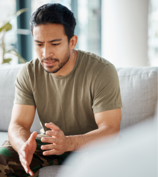 Man with short dark hair and a beard, wearing a green T-shirt, sitting on a couch and looking concerned while talking to someone.