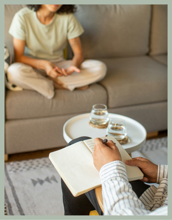 Person in a striped shirt writing in a notebook while sitting on a chair; another person in casual clothing sitting on a sofa in the background; two glasses of water on a tray on a small table.