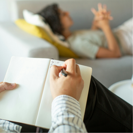 A person writing in a notebook while a woman is lying on a sofa in the background