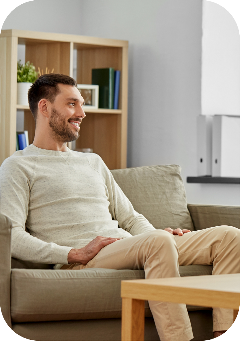 A smiling man sitting on a beige sofa in a modern living room with a wooden bookshelf and white wall behind him.
