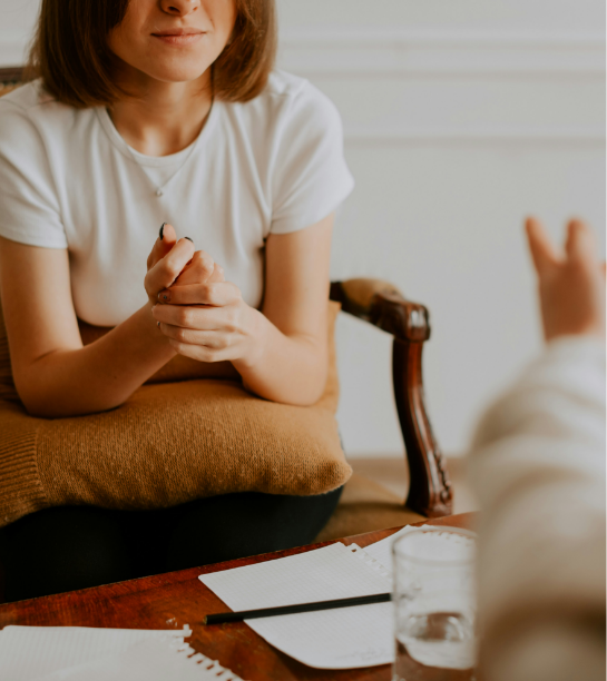 A woman with shoulder-length brown hair sitting at a table, hands clasped, talking to a person whose arm is visible in the foreground.