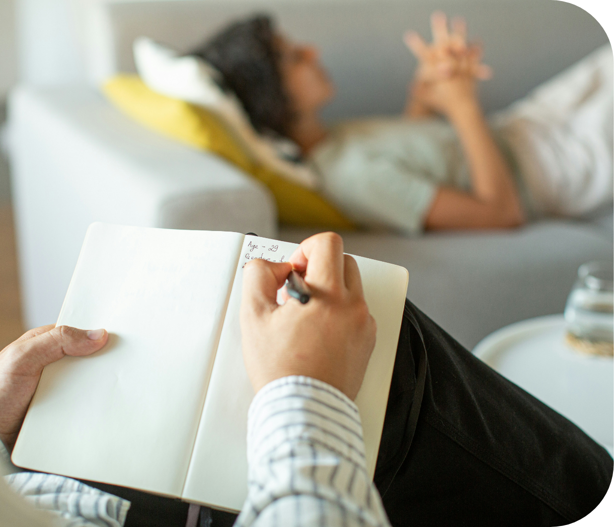 Person sitting on a chair taking notes in a notebook while a woman lies on a sofa in the background, blurred. Water glass on a table nearby.