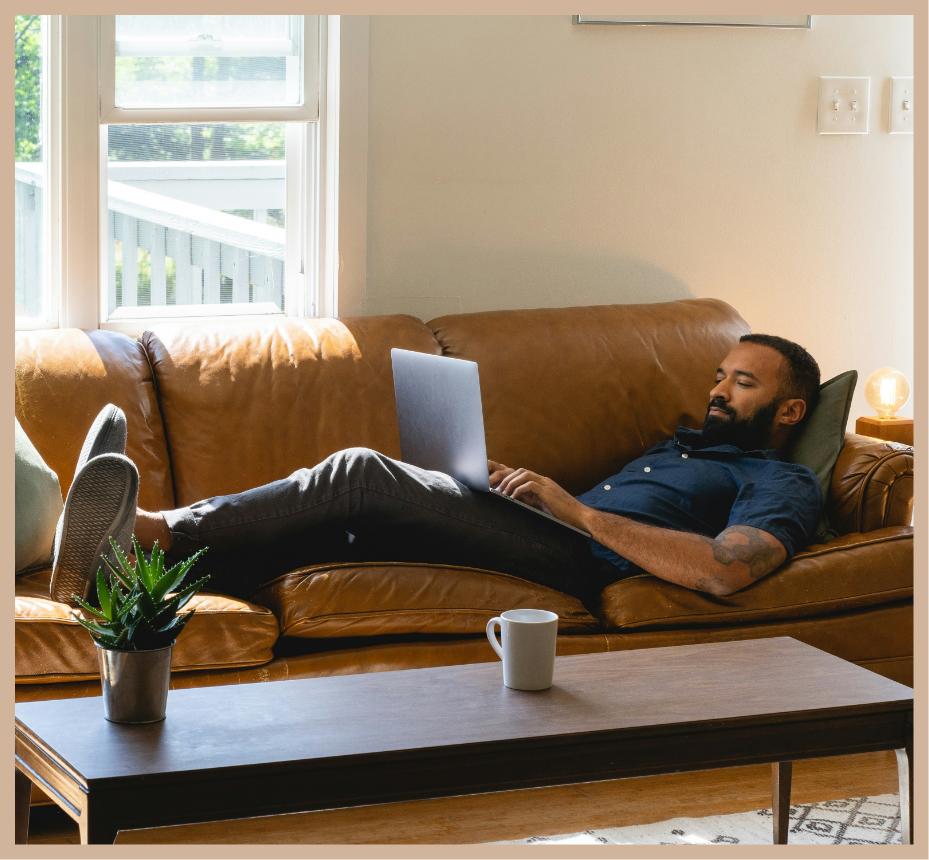 A man lying on a brown leather couch using a laptop.