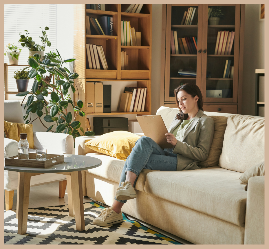 Woman reading a book on a beige sofa in a living room with wooden bookshelves and a large green plant.