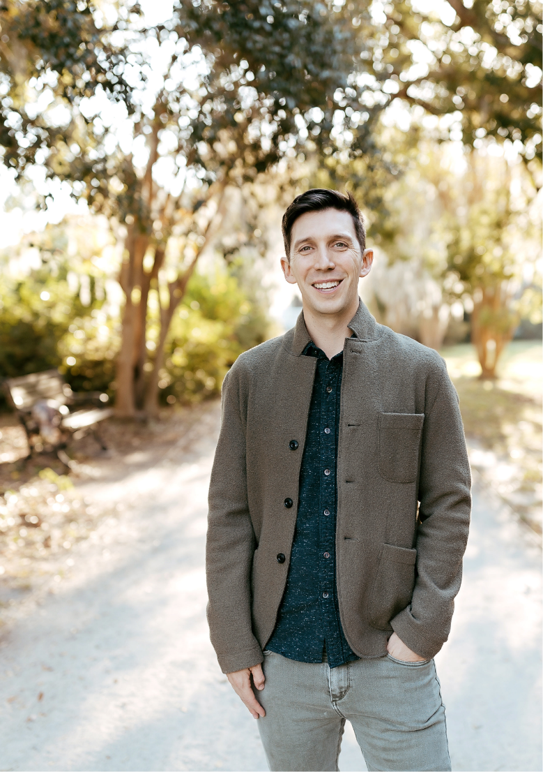 A young man with dark hair, dressed in a grey jacket and dark shirt, standing outdoors on a sunlit path with trees and benches in the background, smiling at the camera.