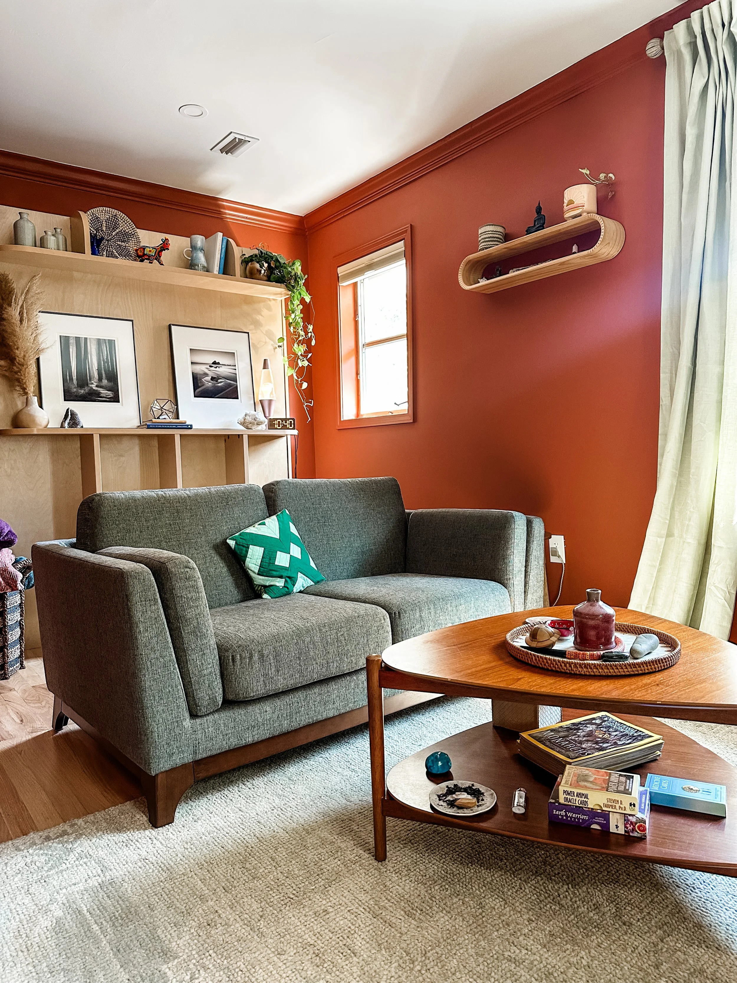 A living room with a green sofa, wooden coffee table, an orange wall, and decor items including framed pictures, a shelf with small objects, and a curtain covering a window.