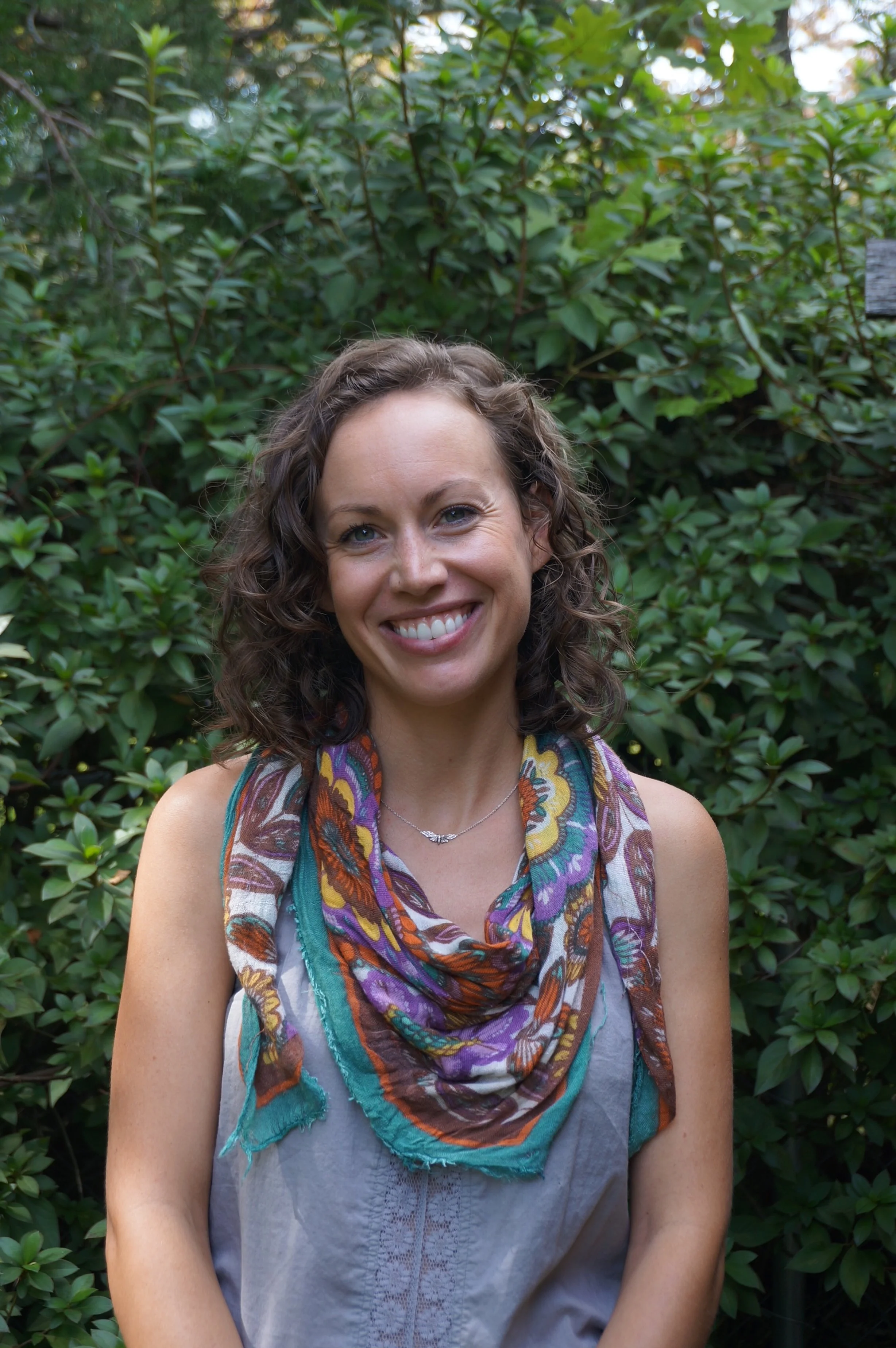 A woman with curly brown hair smiling outdoors, wearing a colorful scarf and a gray sleeveless top, with green leafy bushes in the background.