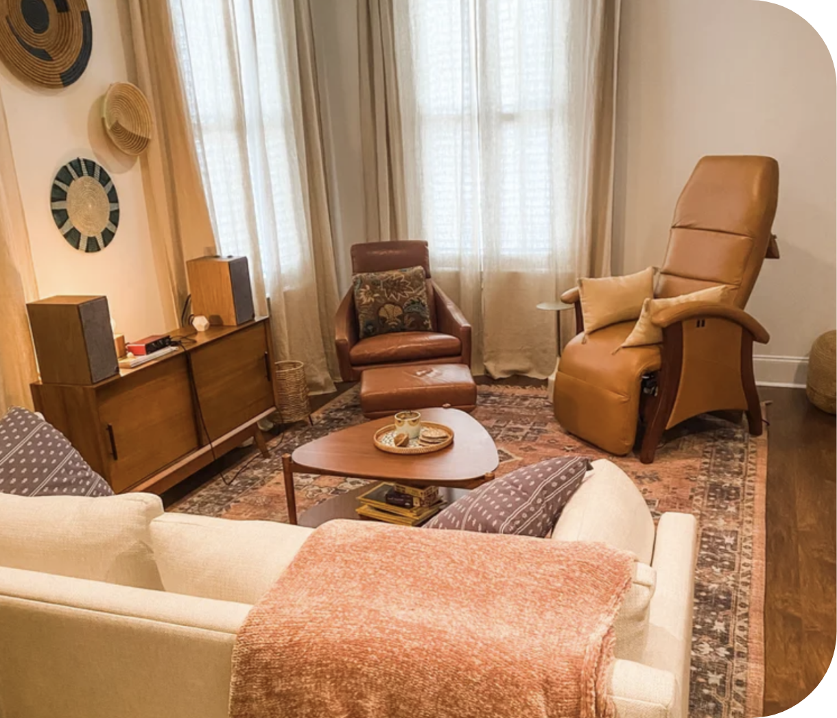 Cozy living room with vintage wooden sideboard, two armchairs, and a sofa with a pink throw blanket, centered around a wooden coffee table holding a mug and snacks, illuminated by natural light from large curtains.